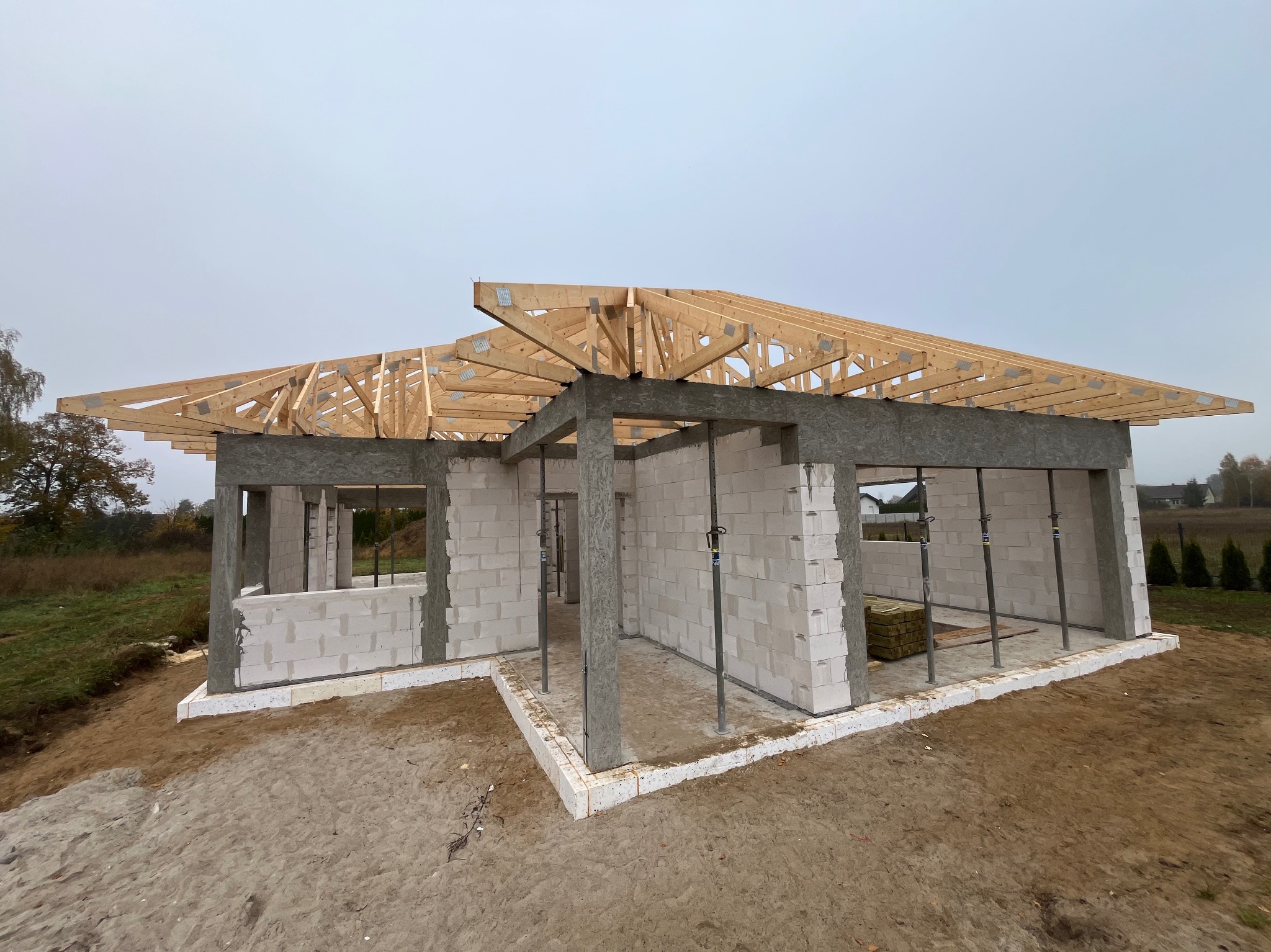 Surowy stan domu jednorodzinnego z białych bloczków, widoczne drewniane elementy konstrukcji dachu, szare słupy i podpora, ziemia wokół budynku.