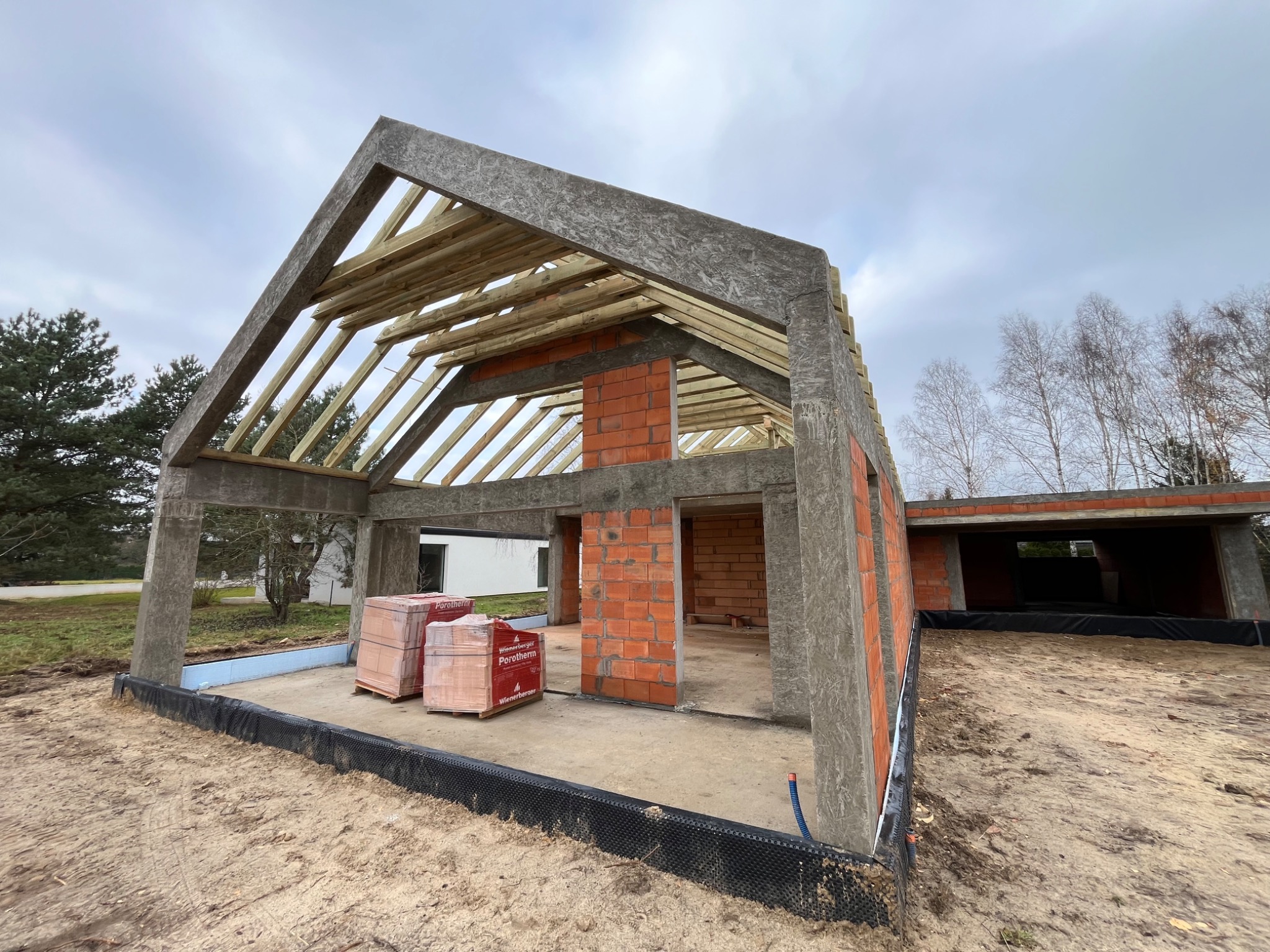 Surowy stan budynku z betonu i cegły Porotherm, widoczne elementy konstrukcyjne dachu z drewna, teren budowy z czarną folią fundamentową i kostkami cegieł na paletach.