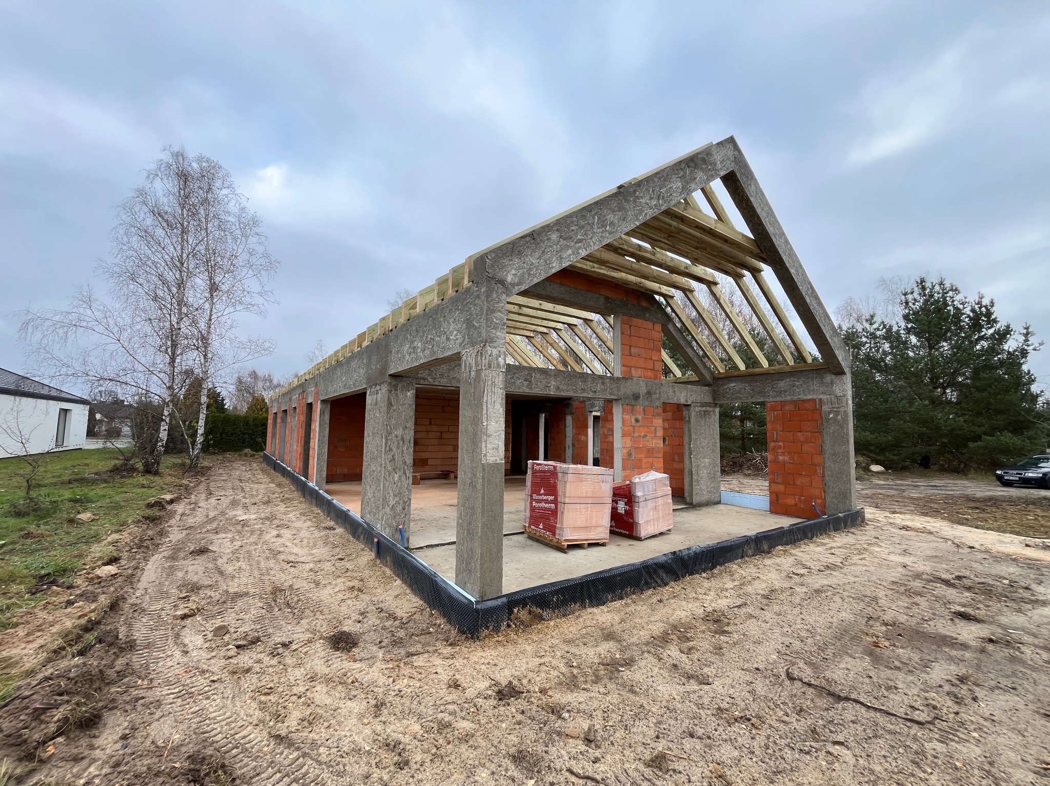 Surowy stan domu murowanego z betonu komórkowego i elementami żelbetowymi, widoczne bloczki Porotherm na paletach, drewniana konstrukcja dachu, izolacja fundamentów, teren budowy z piaskiem.