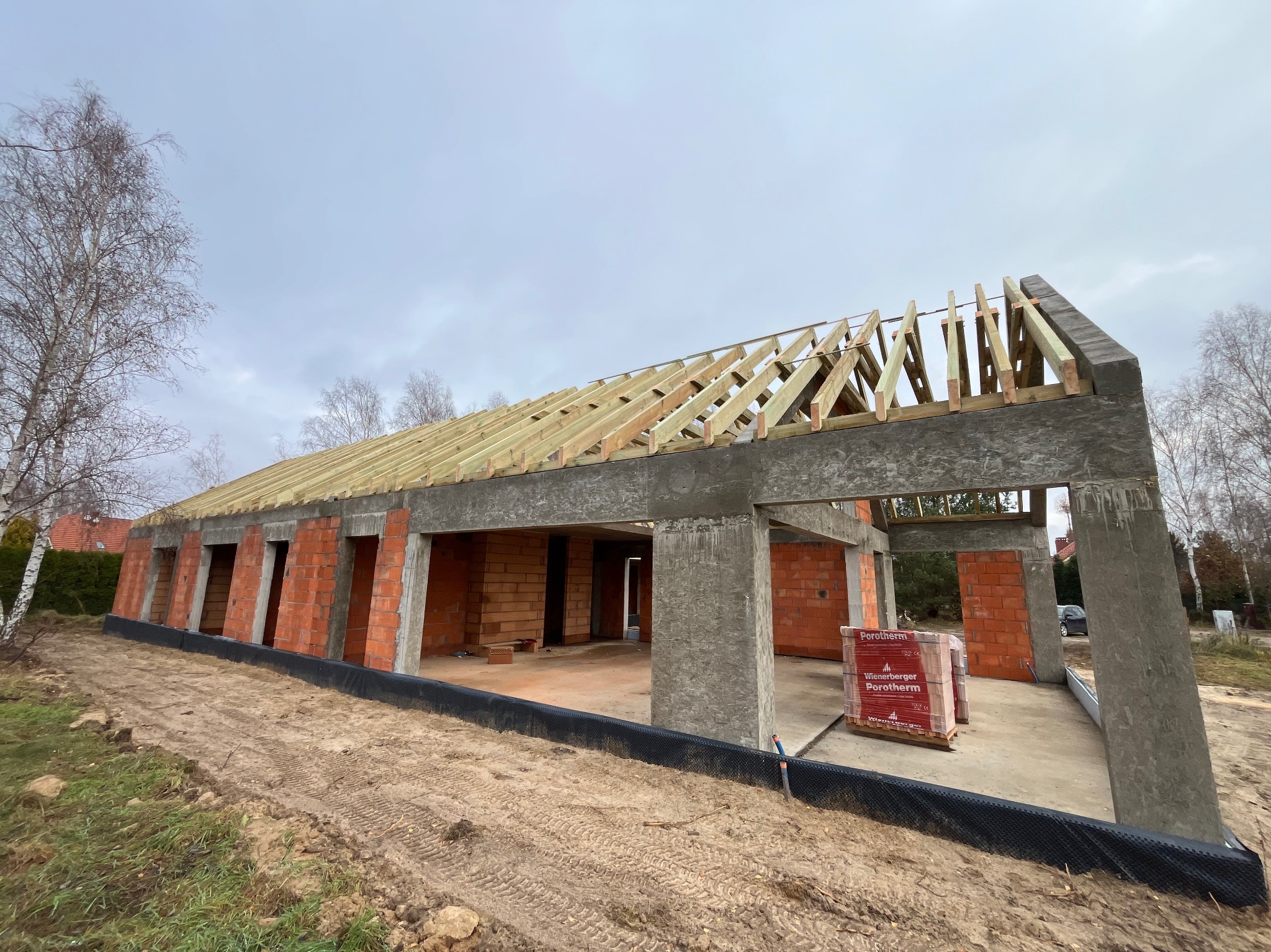 Surowy stan otwarty budynku z czerwonej cegły z betonowymi słupami i drewnianą konstrukcją dachu, paleta cegieł Porotherm na placu budowy