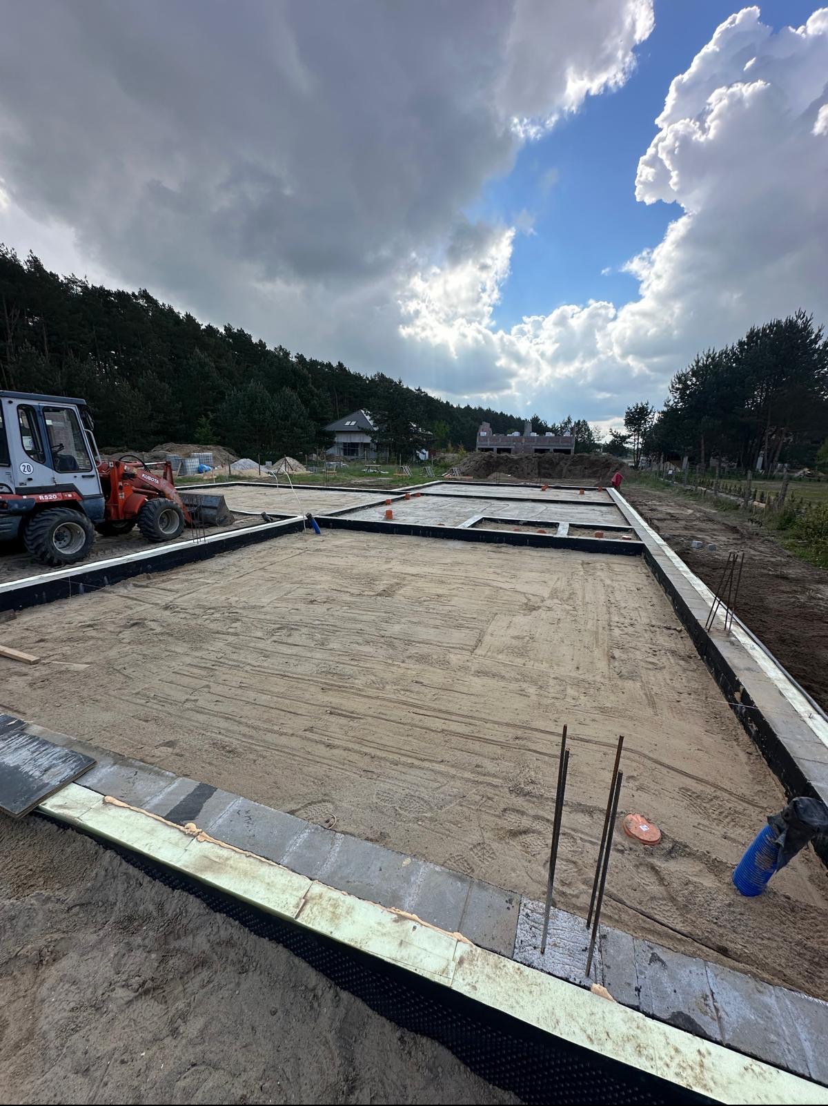 Przygotowanie fundamentów pod budowę domu w Deszcznie: widoczny zarys fundamentów, izolacja przeciwwilgociowa, wystające pręty zbrojeniowe oraz minikoparka na placu budowy w tle.