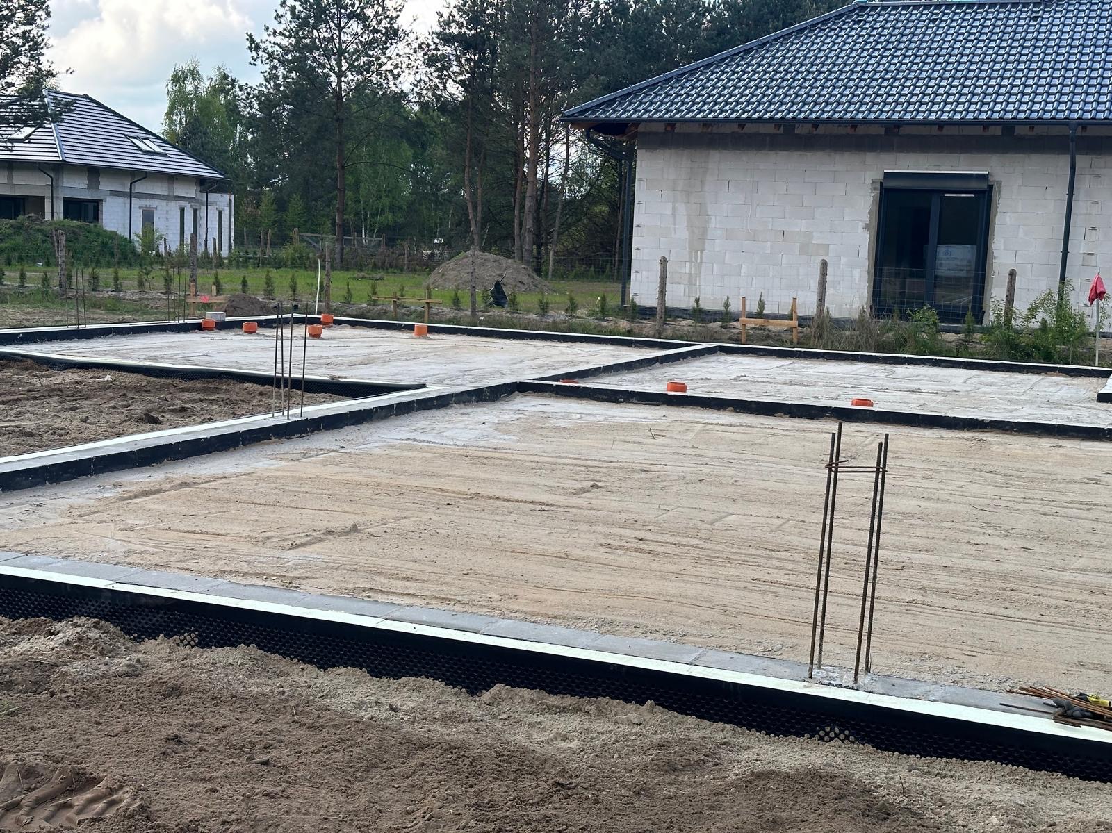 Przygotowany teren pod budowę domu z widoczną izolacją fundamentów, wystającymi prętami zbrojeniowymi i częściowo wylaną płytą fundamentową. W tle widać dwa budynki w stanie surowym otwarte.