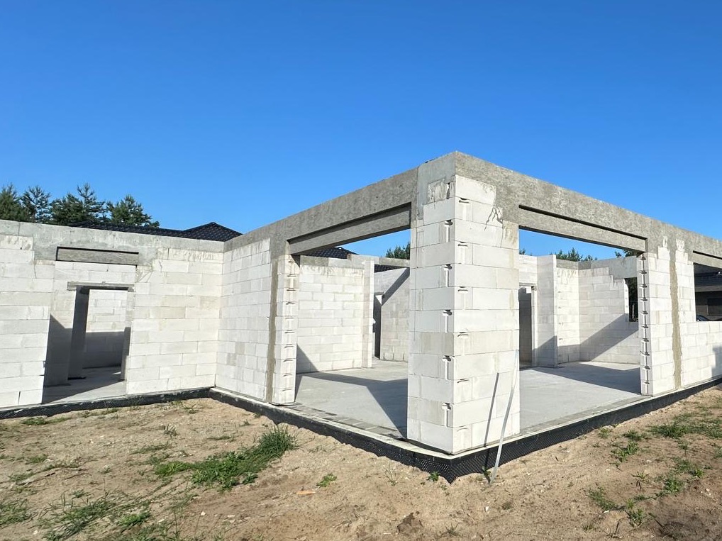 Surowy stan domu z białych bloczków, widoczne ściany nośne i betonowe nadproża, teren wokół budynku częściowo porośnięty trawą, w tle drzewa i fragment dachu sąsiedniego domu, błękitne niebo.