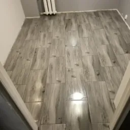 Ułożona podłoga z szarych, prostokątnych płytek imitujących drewno, widoczne fugi i fragment grzejnika. Widok na podłogę zza framugi drzwi.