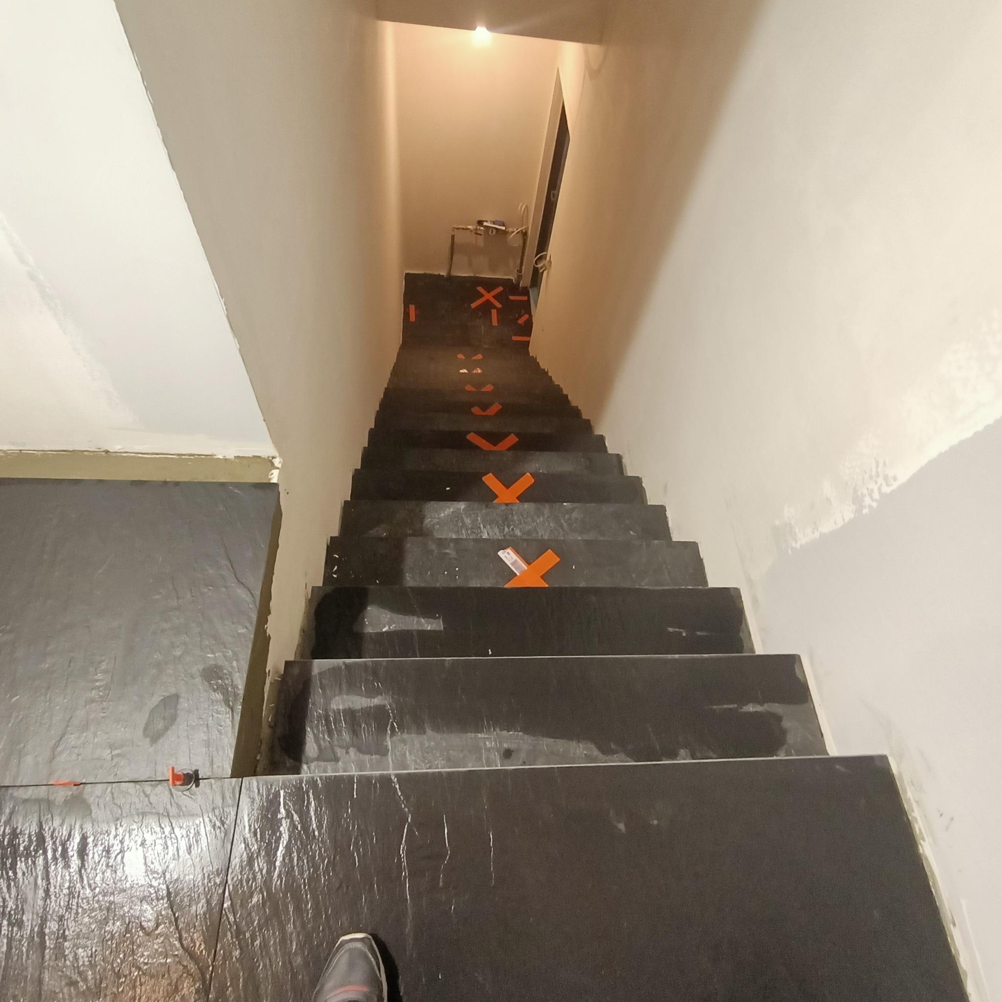 Montaż ciemnych płytek na schodach z użyciem pomarańczowych krzyżyków dystansowych, widok z góry na dół.