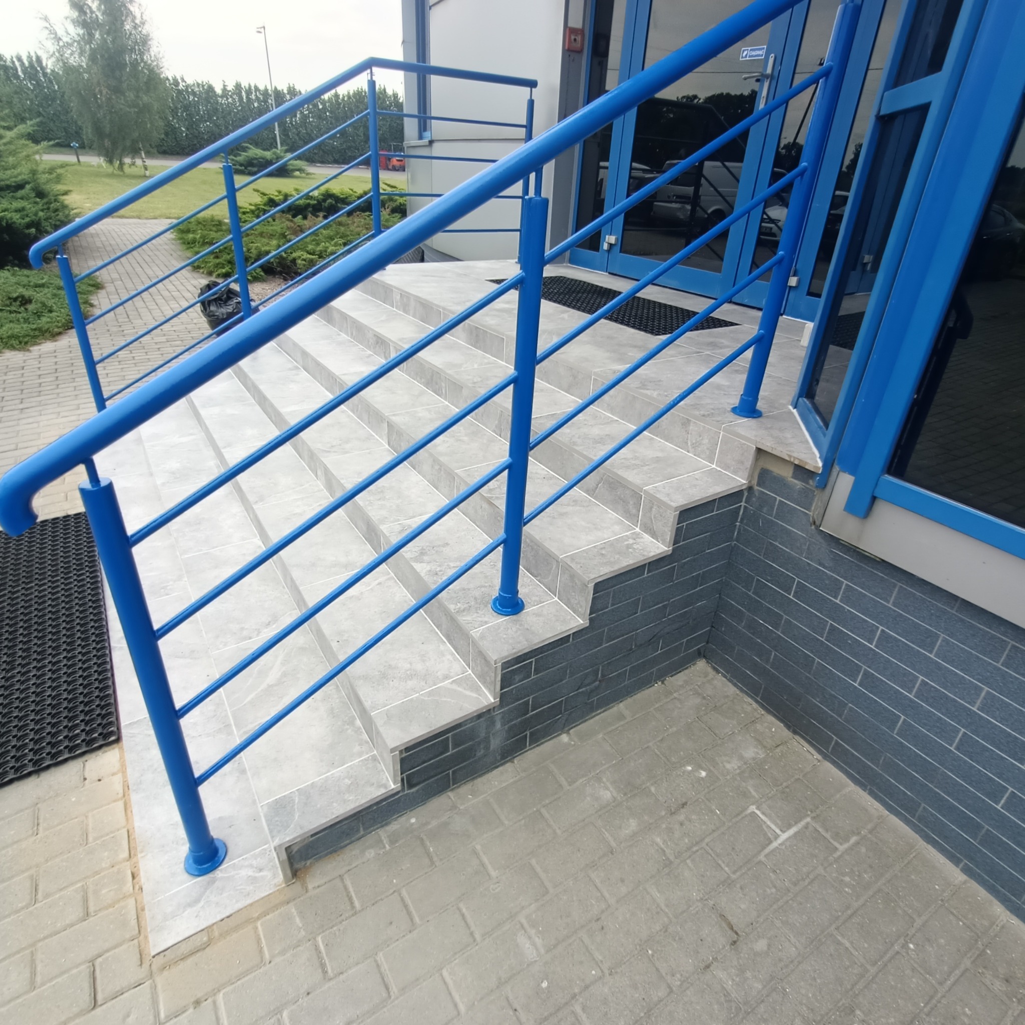 Zewnętrzne schody wejściowe wyłożone szarymi płytkami z niebieską metalową barierką, widok z dołu.