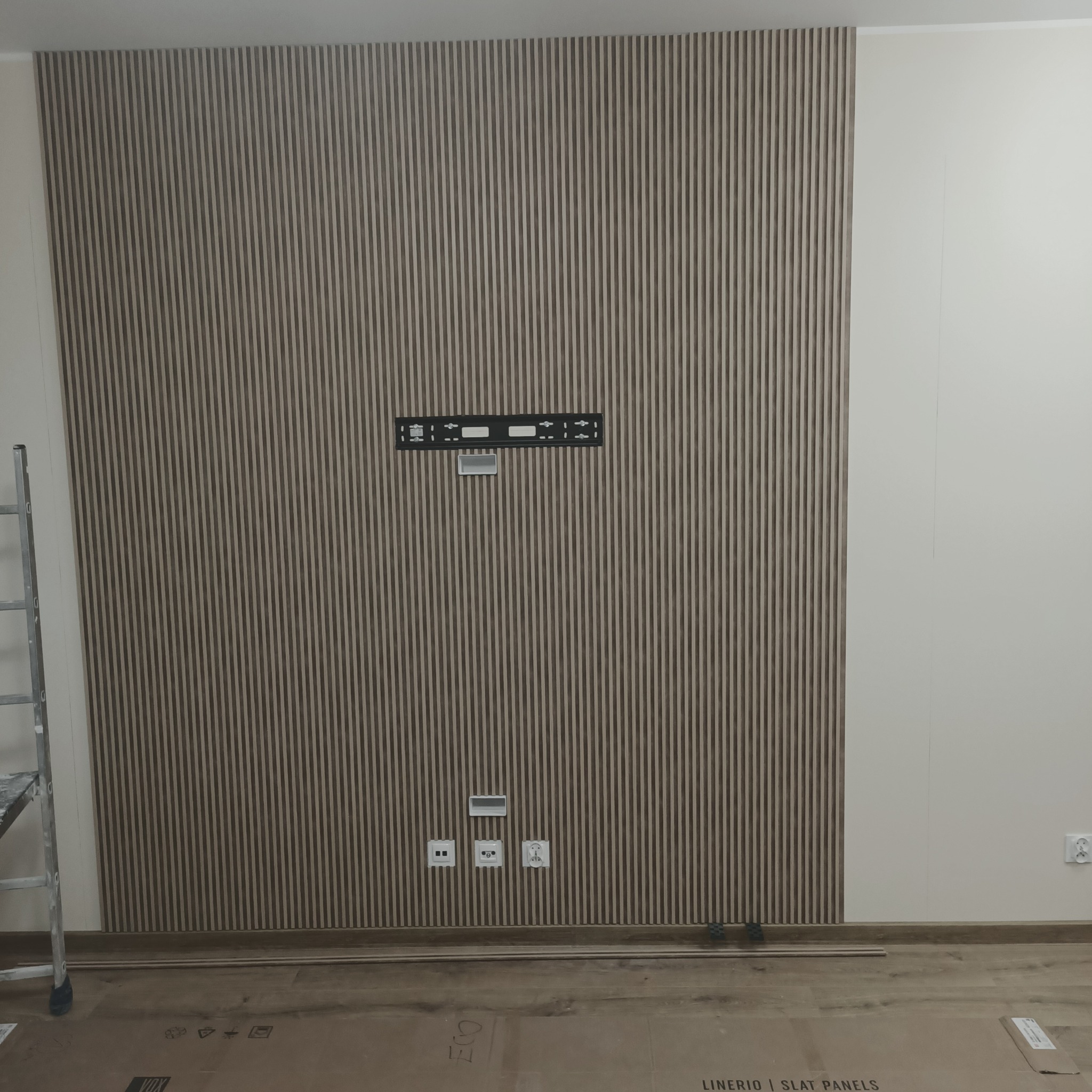 Ściana z zamontowanymi panelami lamelowymi w kolorze drewna, widoczny uchwyt do telewizora, gniazdka elektryczne i fragment drabiny malarskiej.