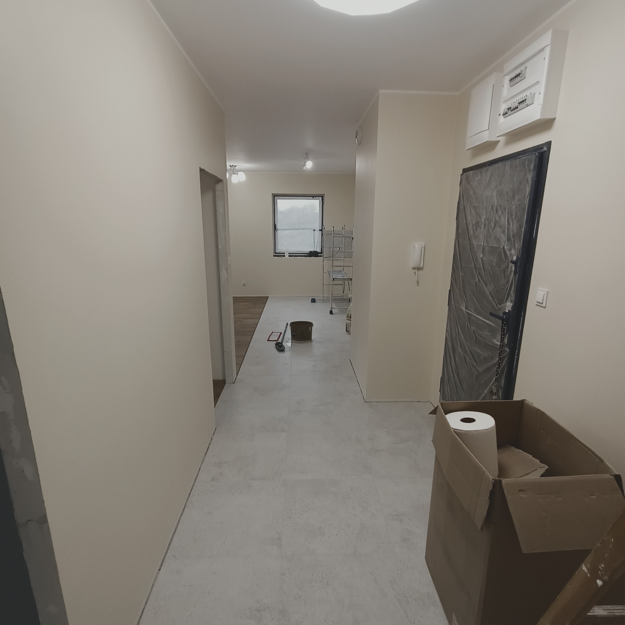 Wnętrze w trakcie prac wykończeniowych: świeżo pomalowane ściany w odcieniach beżu, zabezpieczone folią drzwi, wiadro i narzędzia malarskie na podłodze z jasnych płytek, karton z rolką papieru w rogu.