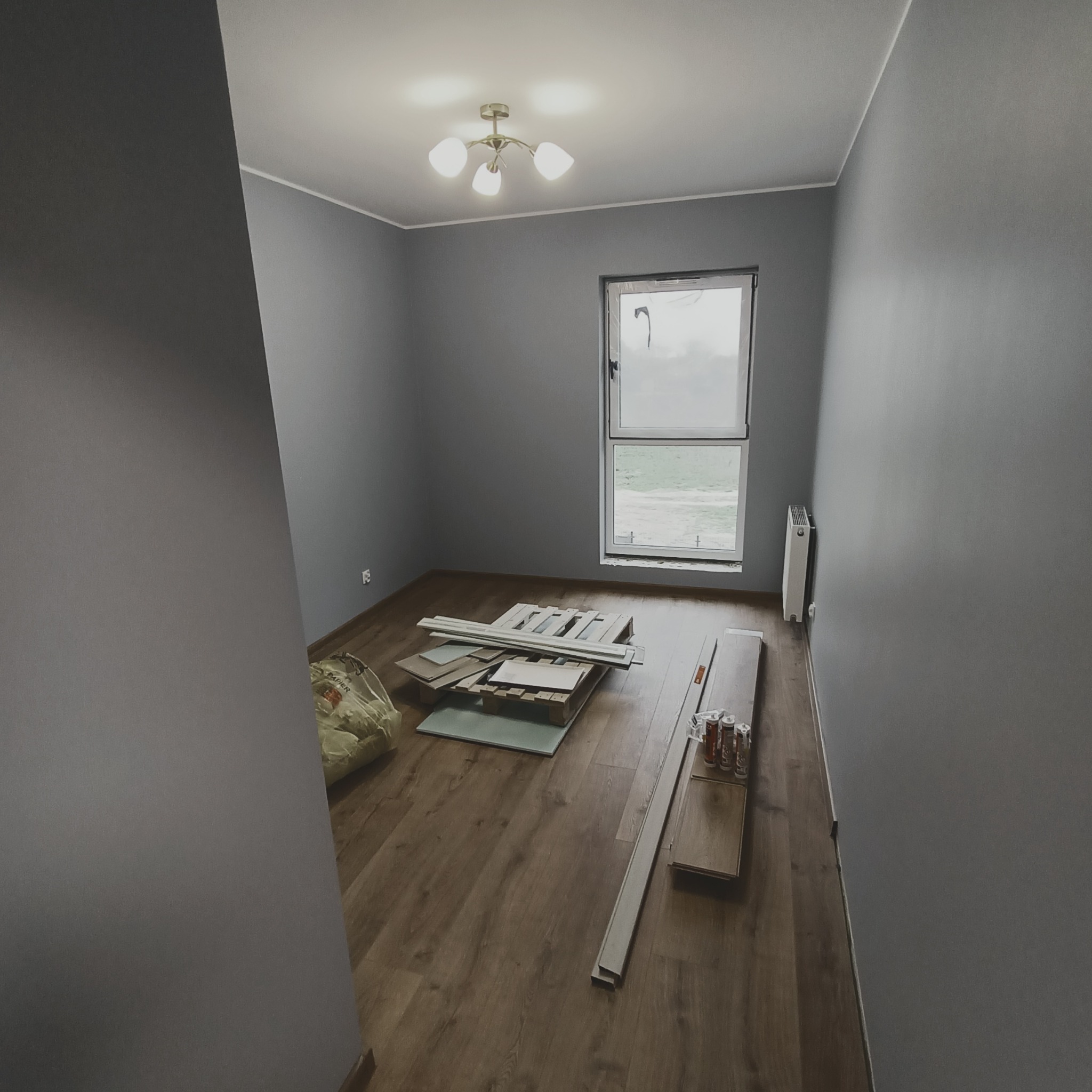 Pokój przed ułożeniem podłogi, widoczne panele podłogowe, listwy przypodłogowe, grzejnik, okno, szare ściany i lampa sufitowa.