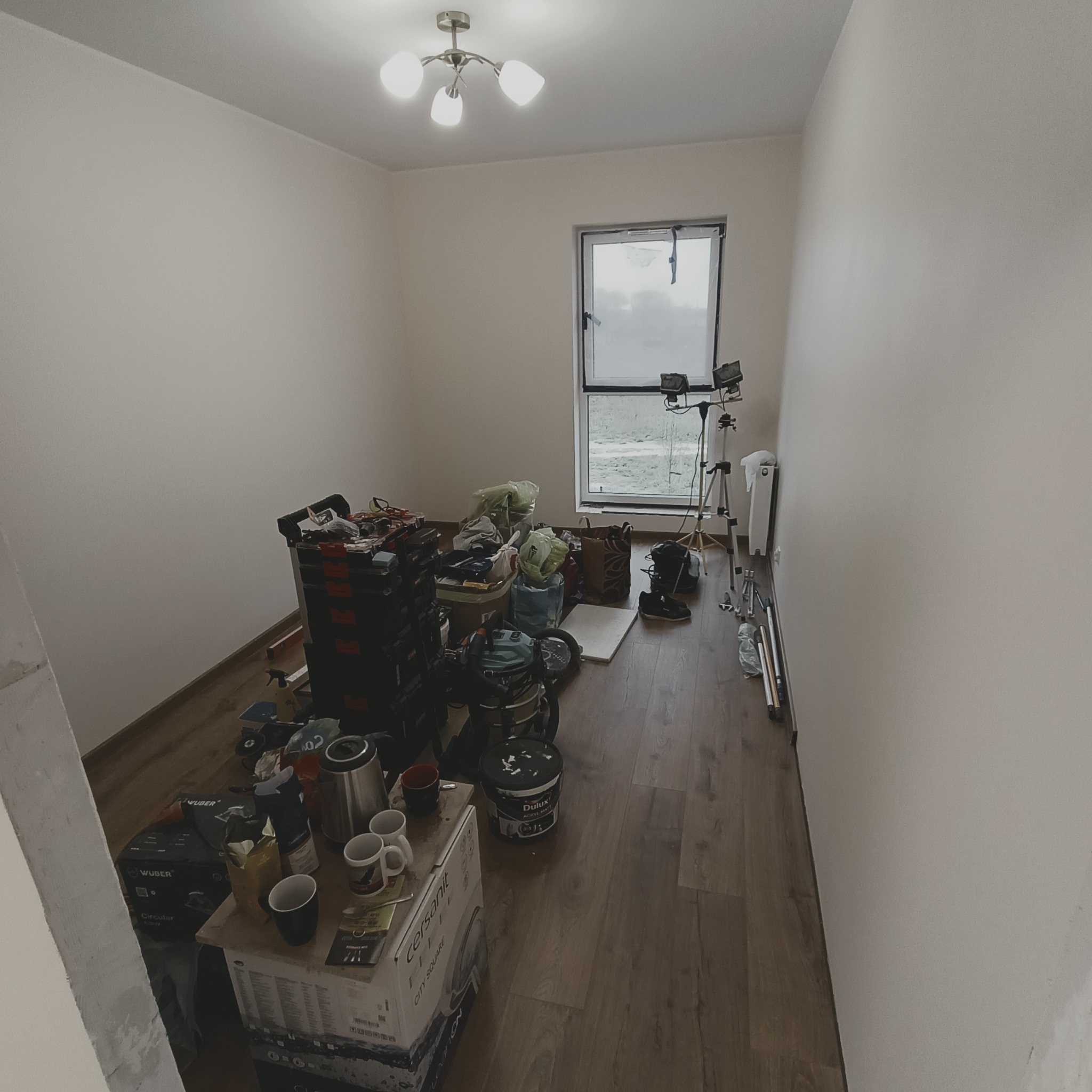 Pokój w trakcie przygotowań do prac wykończeniowych: widoczne narzędzia, pędzle, drabina, wiadro z farbą Dulux, kartony z ceramiką sanitarną, lampa sufitowa i okno z widokiem na zewnątrz.