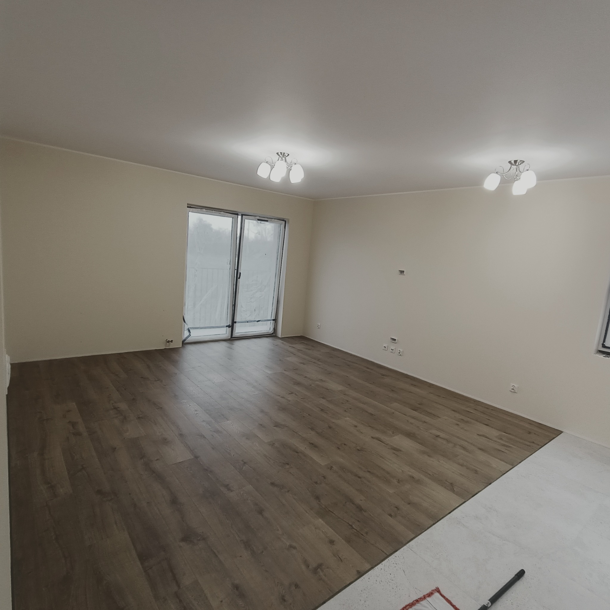 Fragment pokoju z częściowo ułożonymi panelami podłogowymi w odcieniach brązu, widoczne narzędzie do układania paneli i przygotowane podłoże pod dalszą część podłogi.