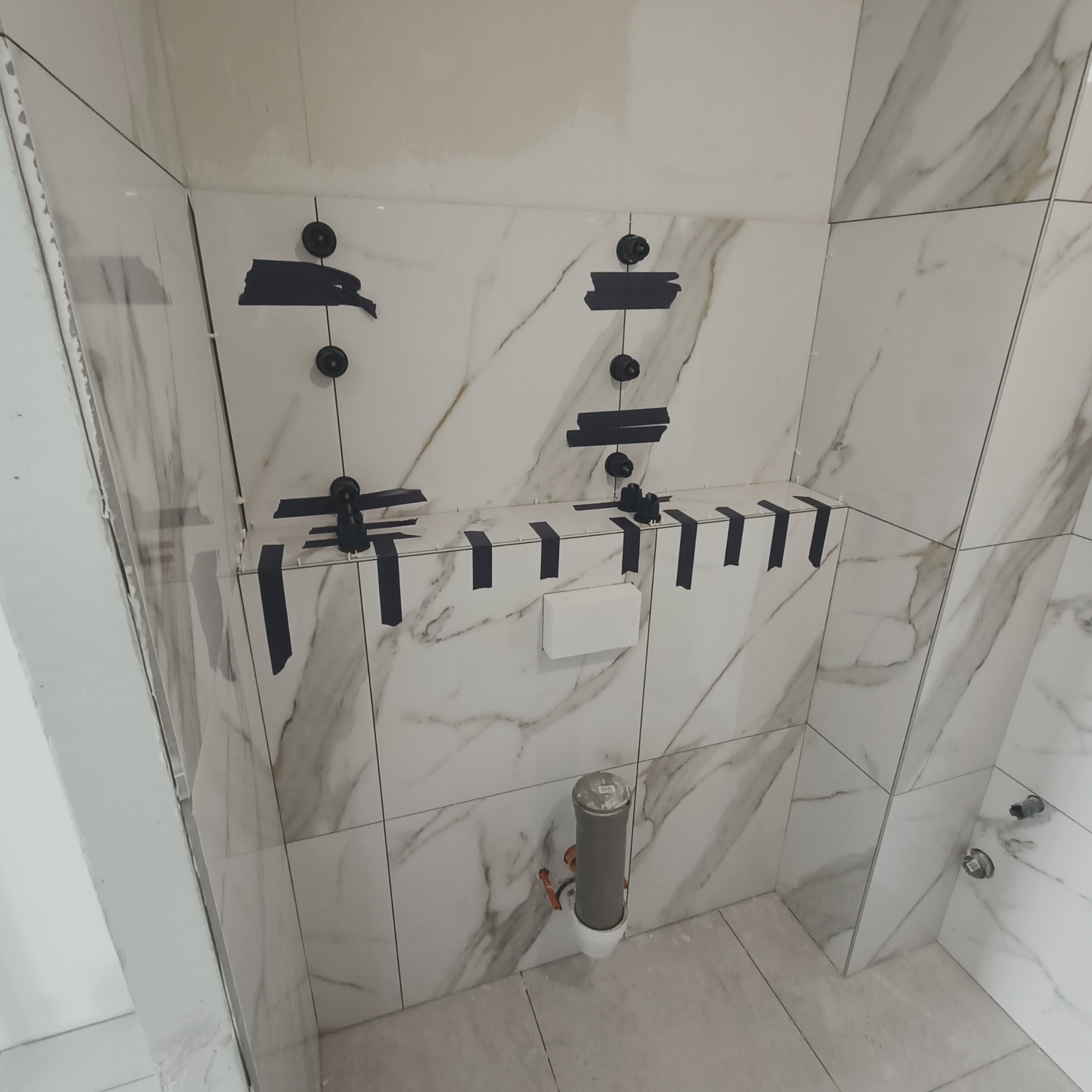 Świeżo ułożone, marmurowe płytki w łazience, z czarnymi klinami i przyssawkami do stabilizacji, widoczny otwór na odpływ w podłodze.