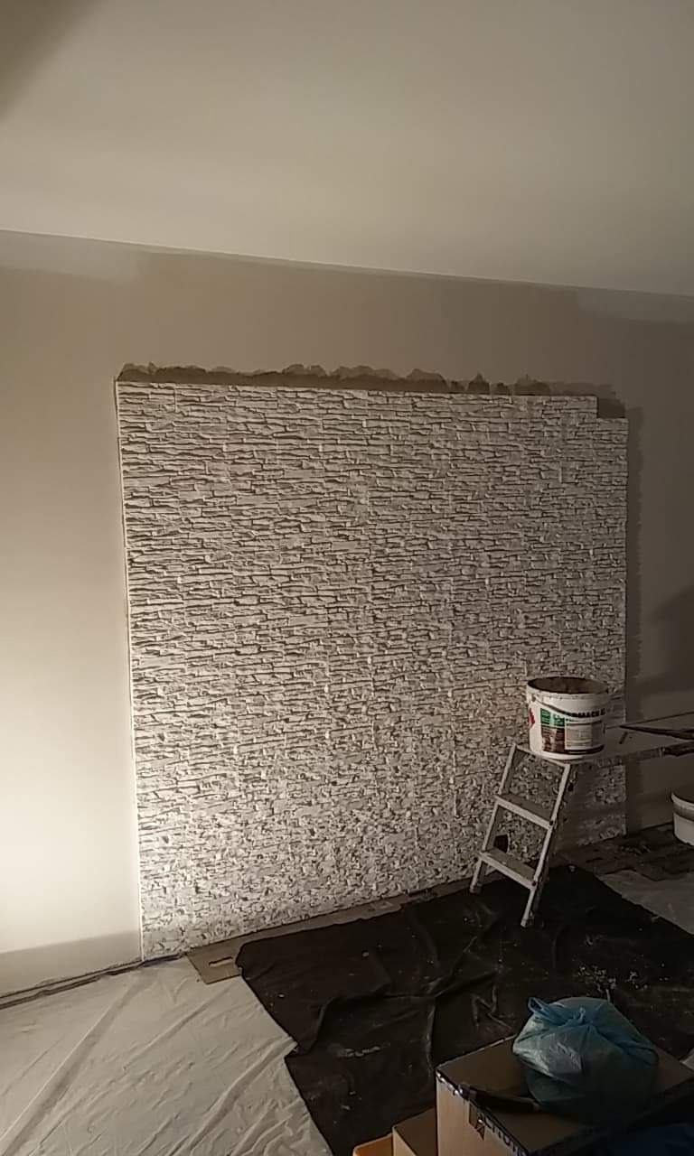 Montaż dekoracyjnej ściany z imitacji kamienia w pomieszczeniu, widoczna drabina malarska i wiadro z klejem, podłoga zabezpieczona folią malarską.