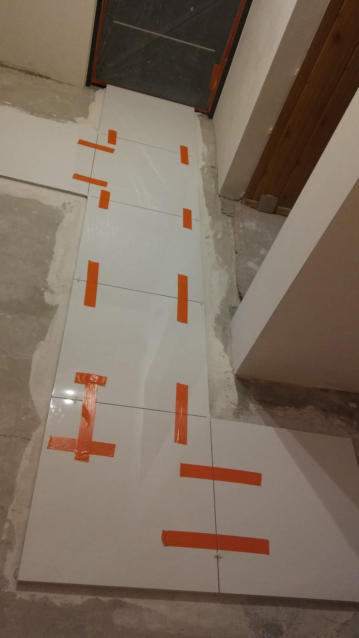 Świeżo ułożone, duże, białe płytki na ścianie, zabezpieczone pomarańczową taśmą malarską i krzyżykami dystansowymi, widoczne surowe wykończenie ścian i sufitu.