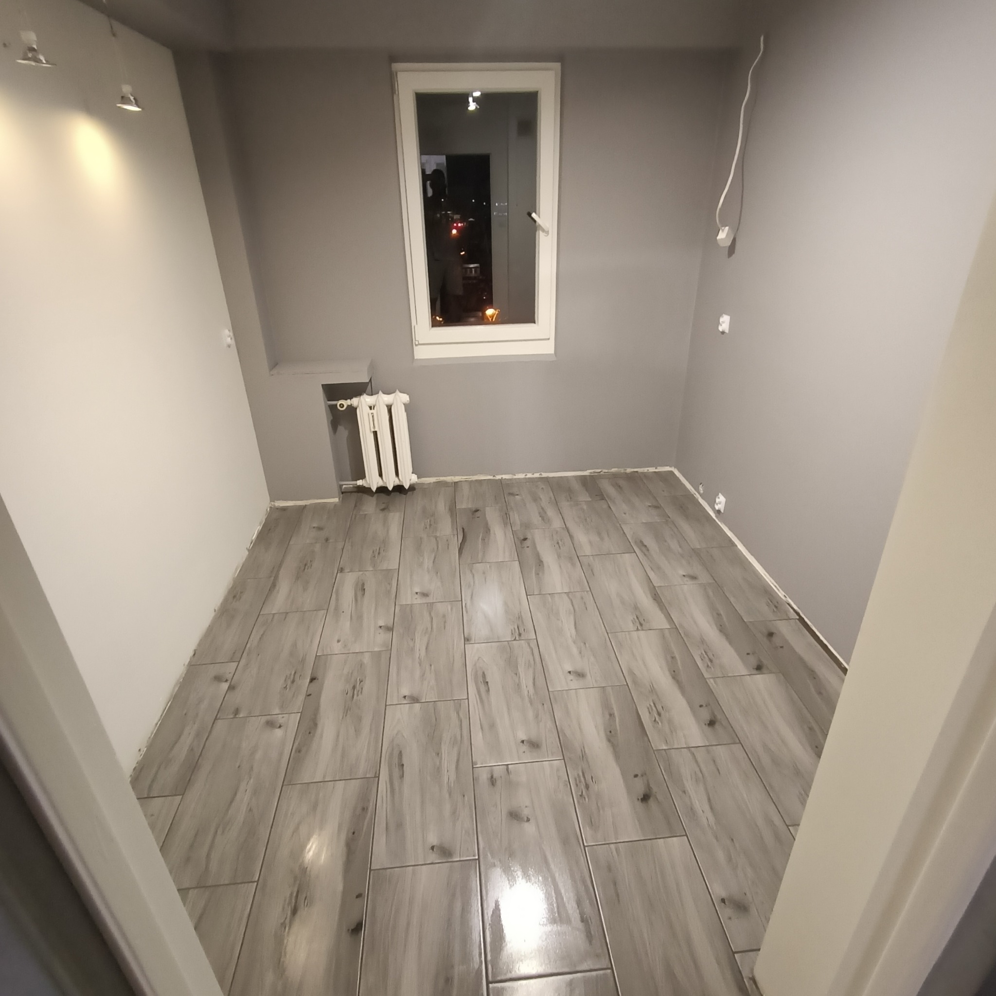 Puste pomieszczenie z nową podłogą z płytek imitujących drewno, szare ściany, białe okno z widokiem na nocne miasto, biały grzejnik i wystające kable elektryczne.