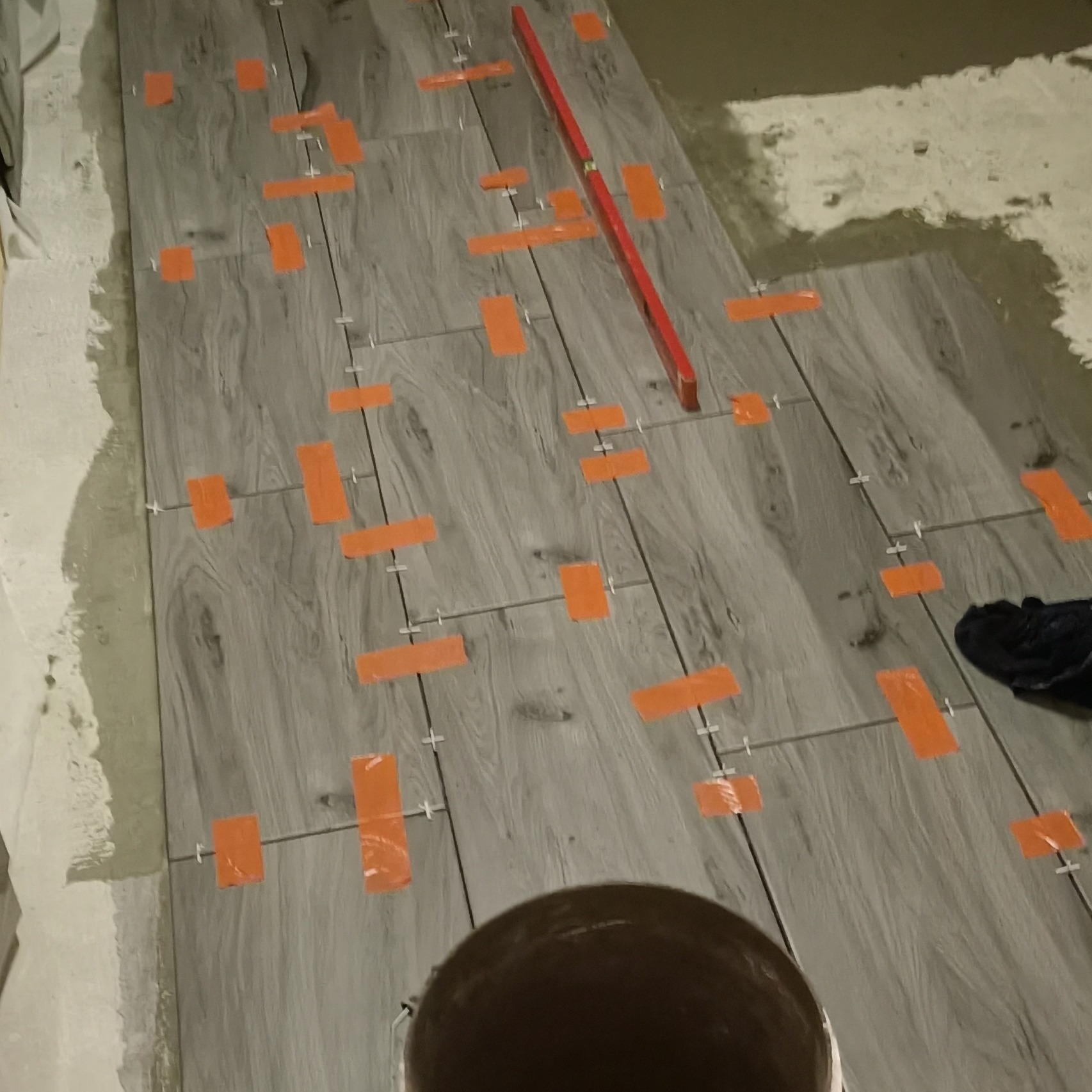 Układanie szarych płytek imitujących drewno na podłodze, z użyciem poziomicy, krzyżyków dystansowych i pomarańczowej taśmy maskującej.