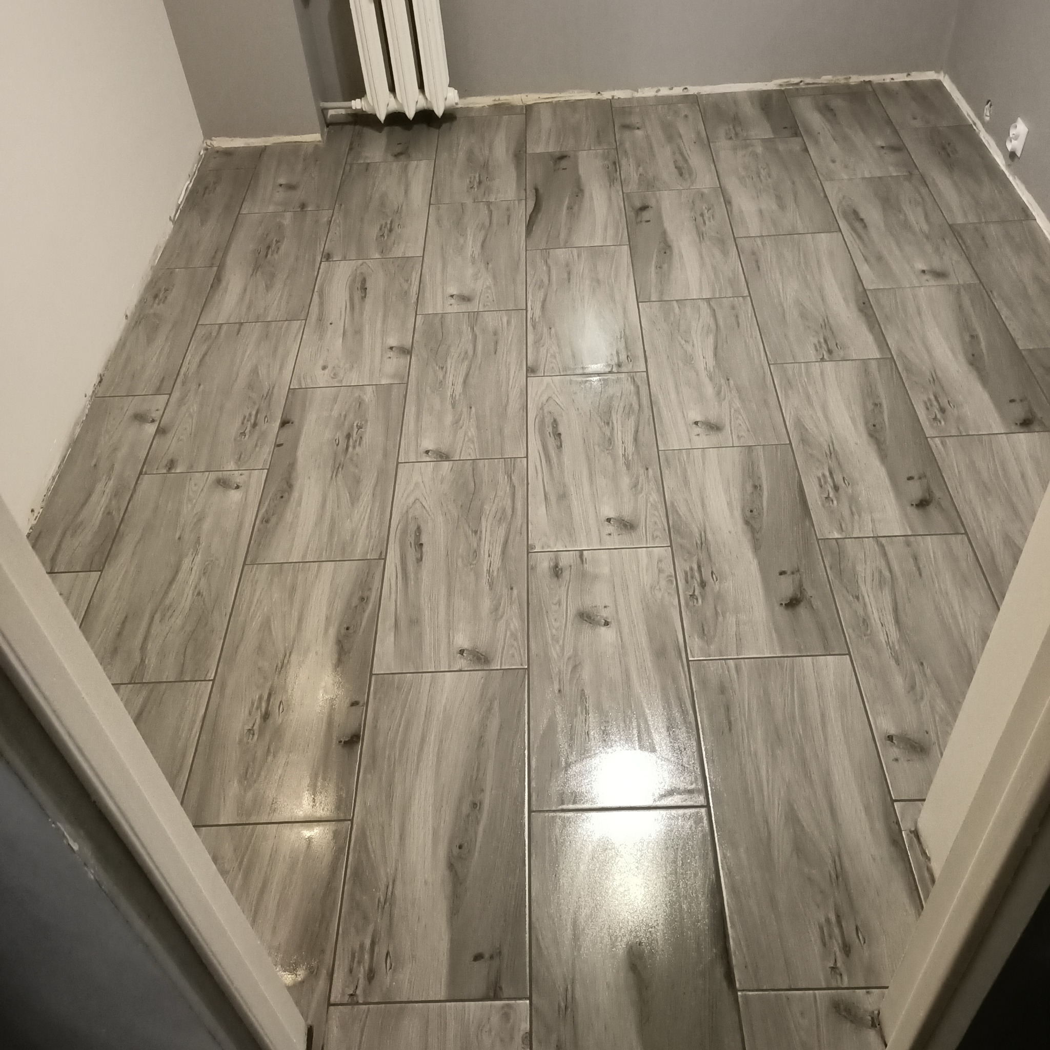 Ułożona podłoga z szarych, prostokątnych płytek imitujących drewno, widoczne fugi i fragment grzejnika. Widok na podłogę zza framugi drzwi.