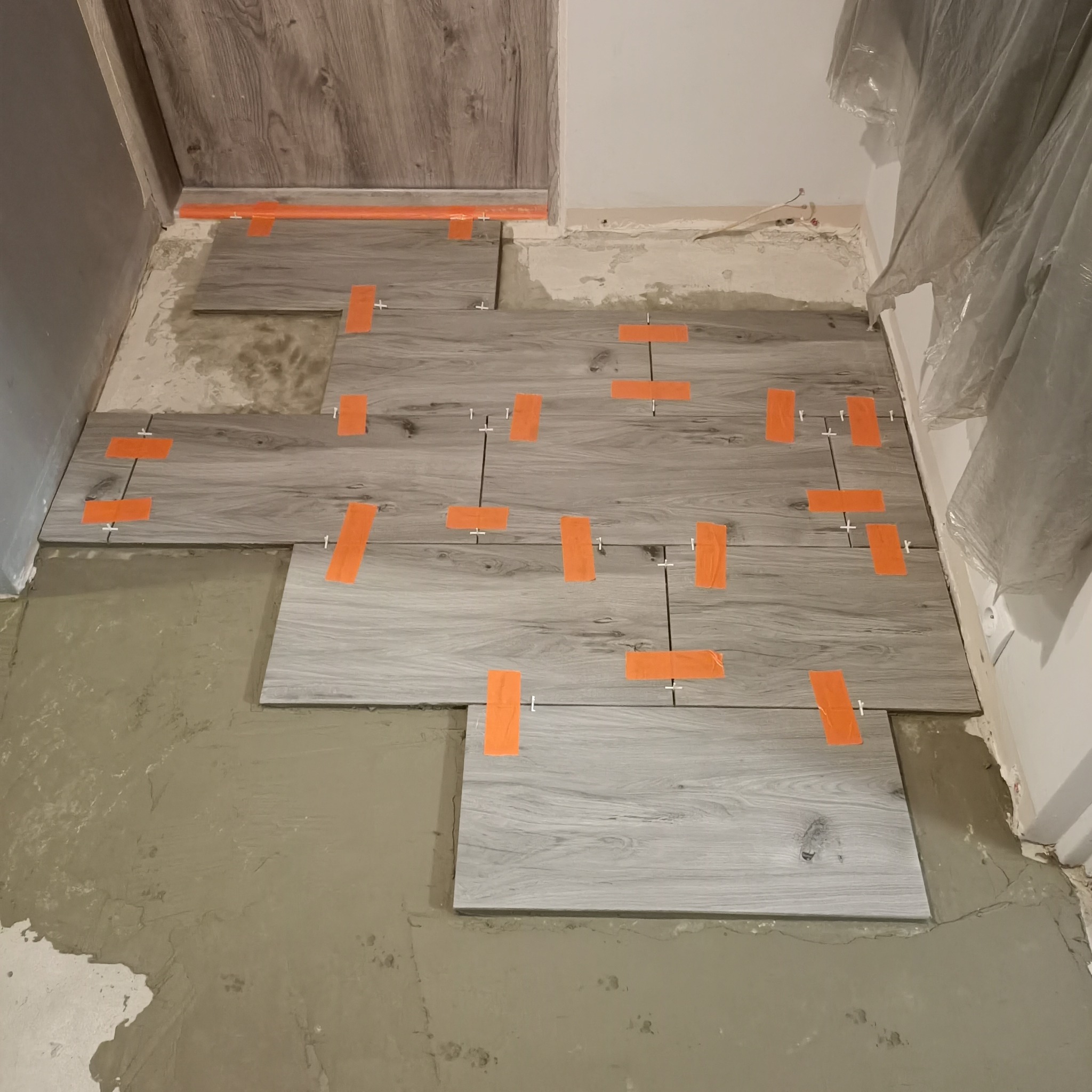Fragment podłogi w trakcie układania płytek imitujących drewno, z pomarańczowymi paskami taśmy i krzyżykami dystansowymi, widoczna warstwa zaprawy klejowej.