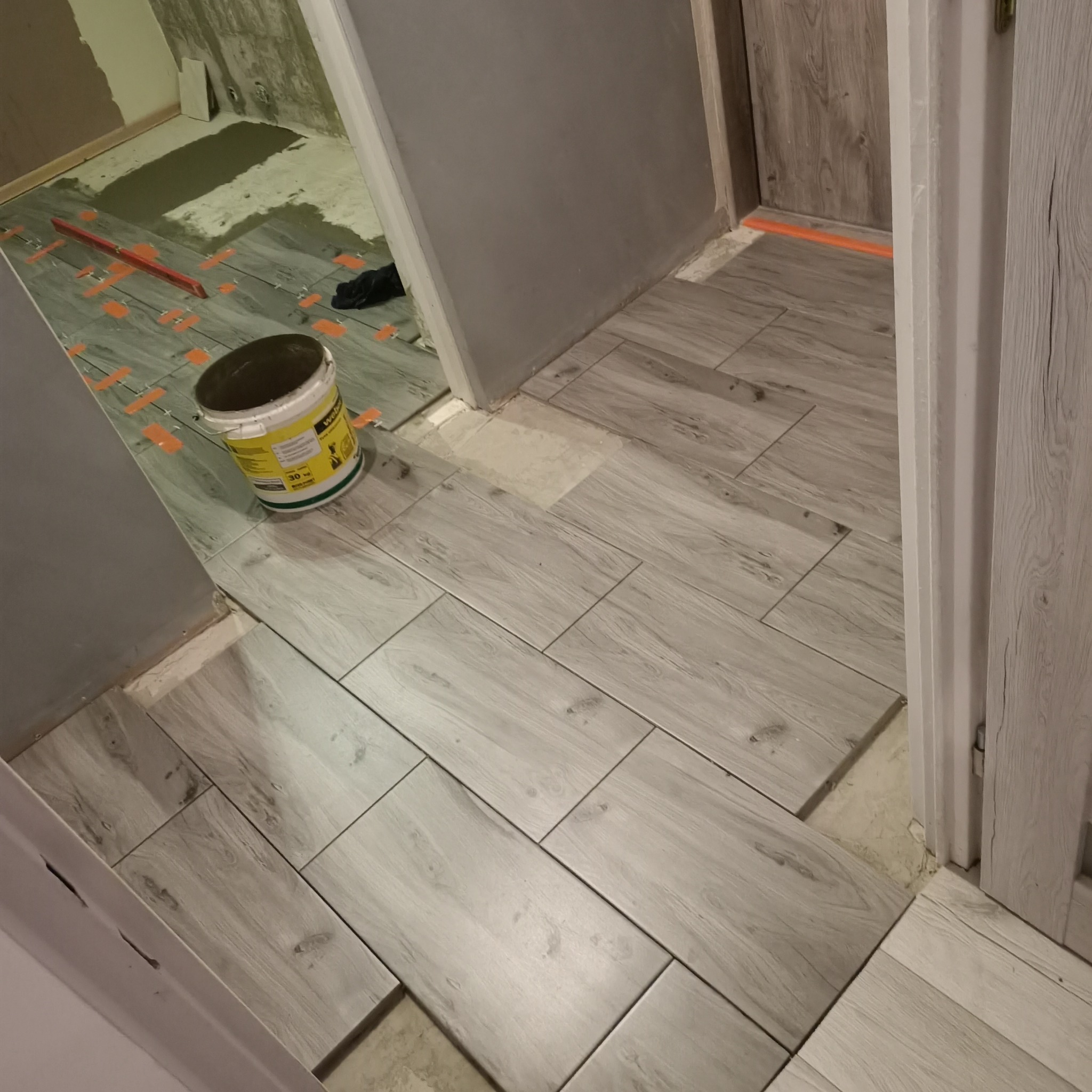 Układanie podłogi z płytek imitujących drewno w trakcie prac, widoczny pojemnik z klejem, poziomica i krzyżyki dystansowe.