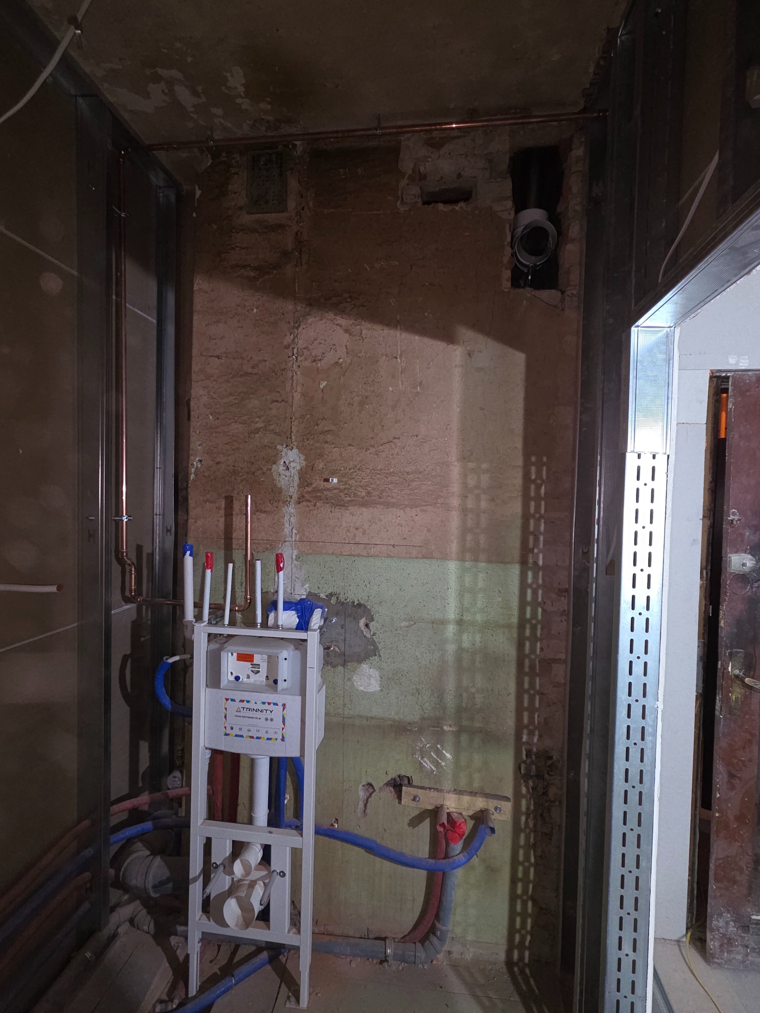 Biały stelaż podtynkowy z instalacją wodną na tle surowej ściany w trakcie remontu. Miedziane rury, niebieskie i białe węże. Widoczne profile konstrukcyjne.