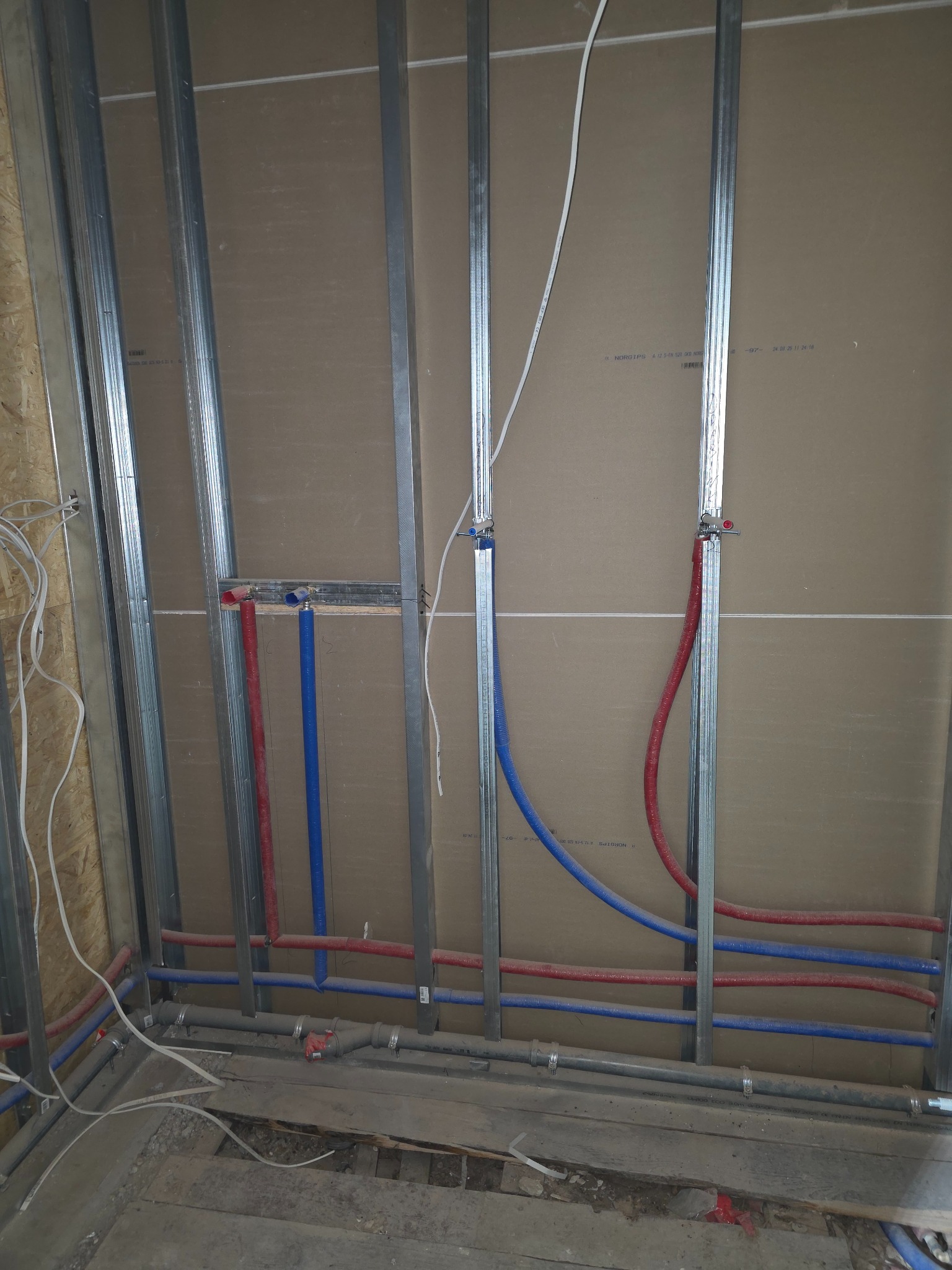 Instalacja wodna w trakcie montażu: czerwone i niebieskie rury prowadzone wzdłuż metalowych profili na tle płyty gipsowej. Widoczne połączenia i elementy mocujące.