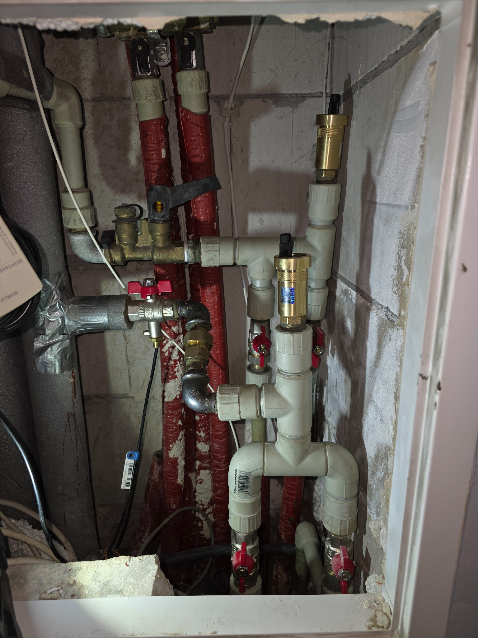 Złożona instalacja wodna z rur PCV i miedzianych, zaworów z czerwonymi pokrętłami i izolowanych rur wnęce. Widoczne elementy montażowe i przyłącza.