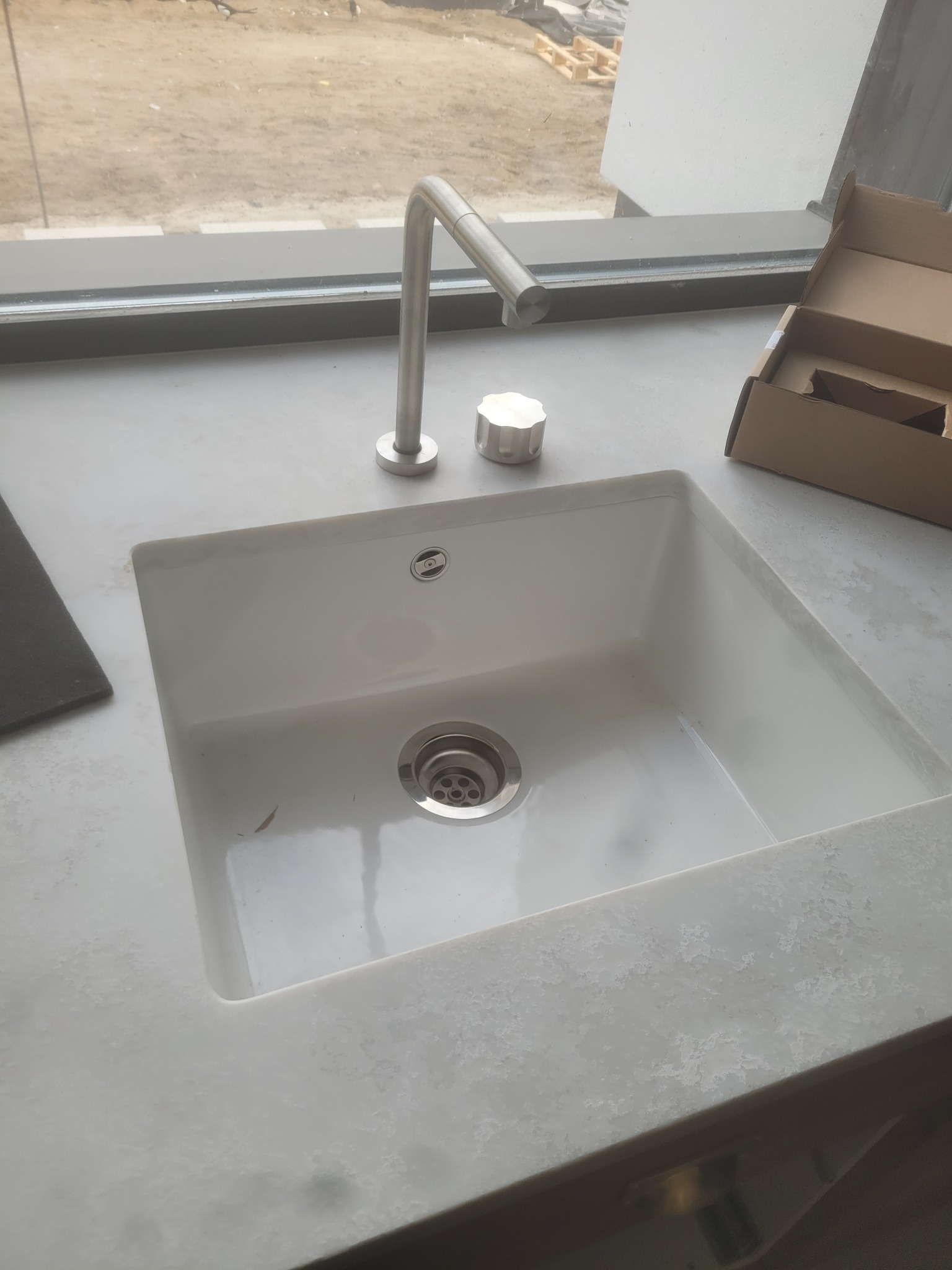 Biała, prostokątna umywalka z chromowaną baterią i korkiem, zamontowana w blacie o jasnej, marmurkowej fakturze, z oknem w tle i kartonowym pudełkiem obok.