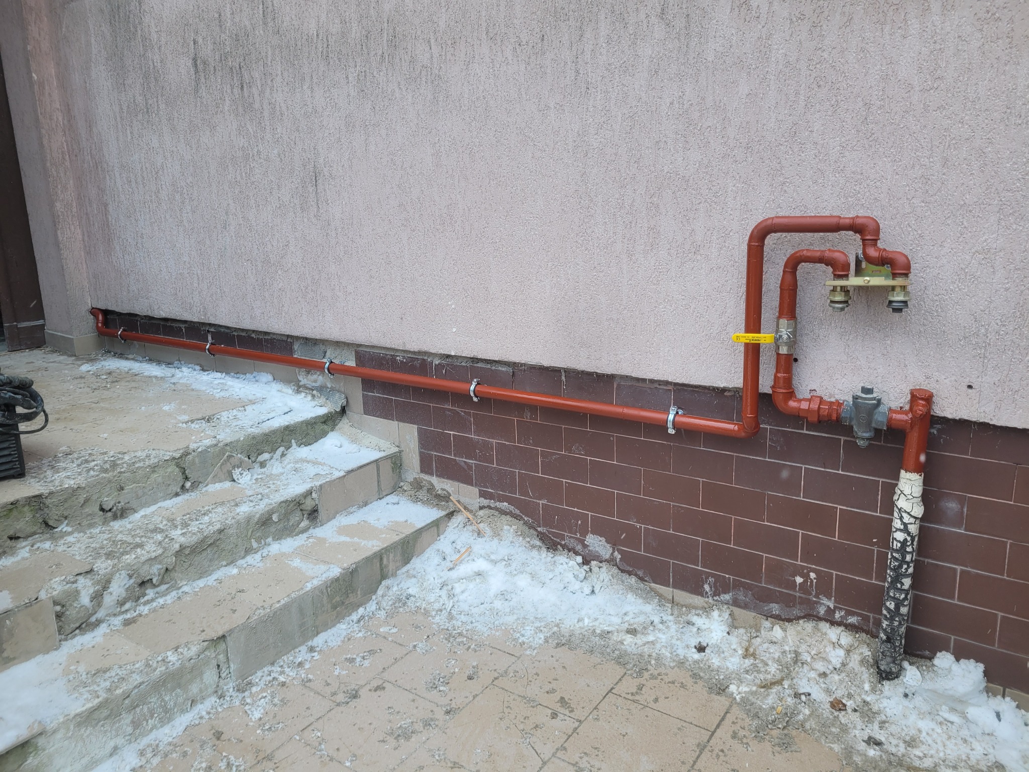 Czerwona instalacja gazowa na zewnątrz budynku, przymocowana do ściany z cegieł i jasnego tynku. Widoczne elementy montażowe i fragment schodów pokrytych śniegiem.
