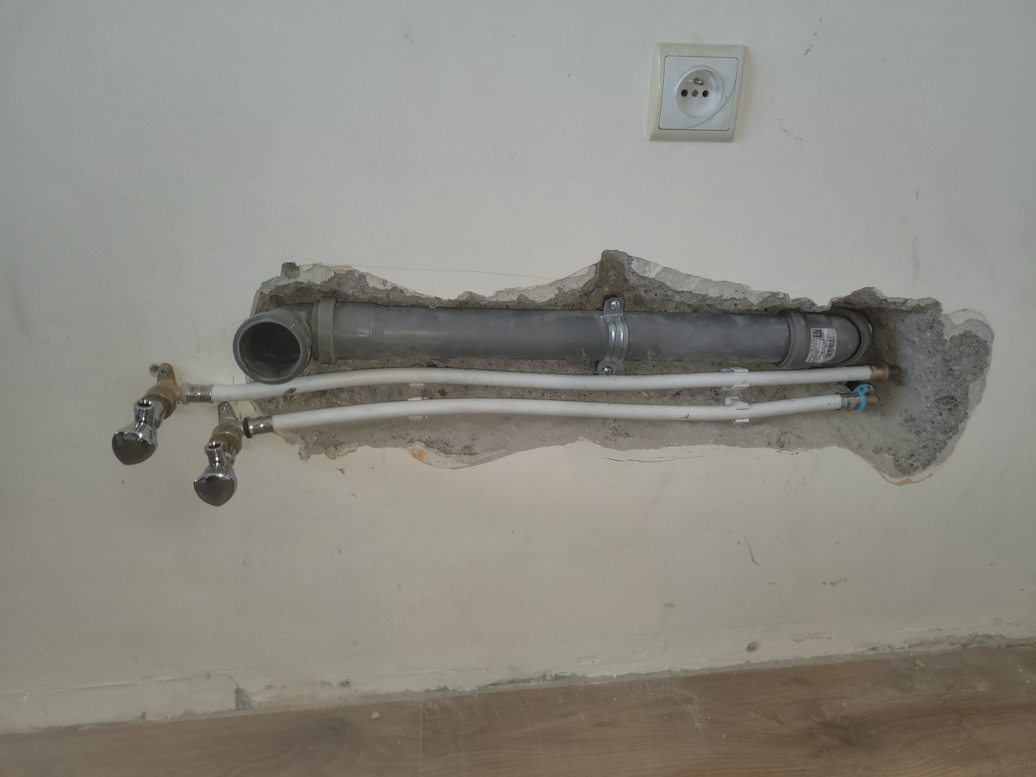 Szare rury kanalizacyjne i białe rury wodne wystające ze ściany, z widocznym gniazdkiem elektrycznym powyżej. Fragment instalacji w trakcie prac.