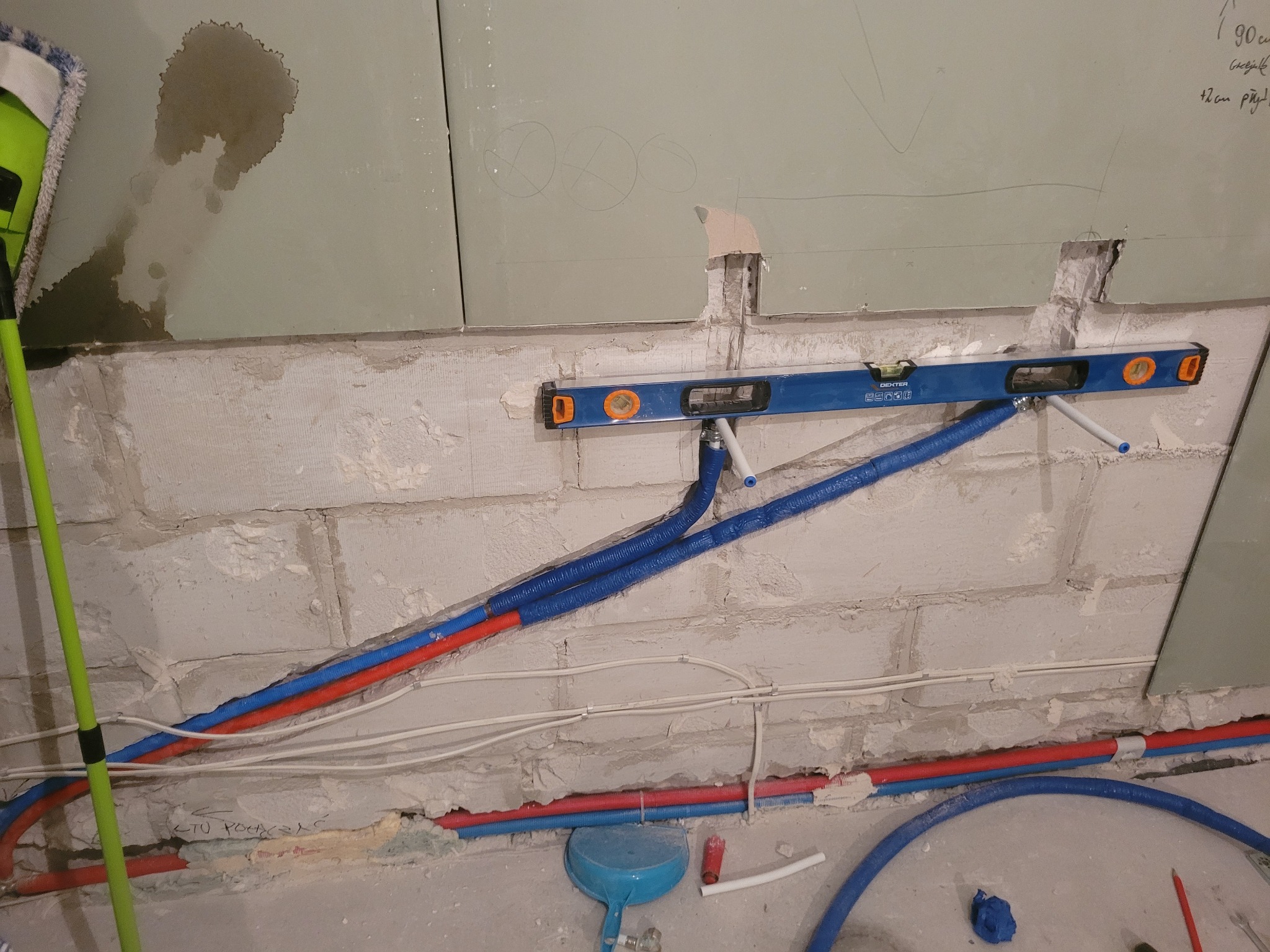Instalacja rur wodnych w ścianie z pustaków, poziomica Dexter, niebieskie rury w izolacji, czerwone rury, białe kable, ściana z płyt kartonowo-gipsowych z oznaczeniami.