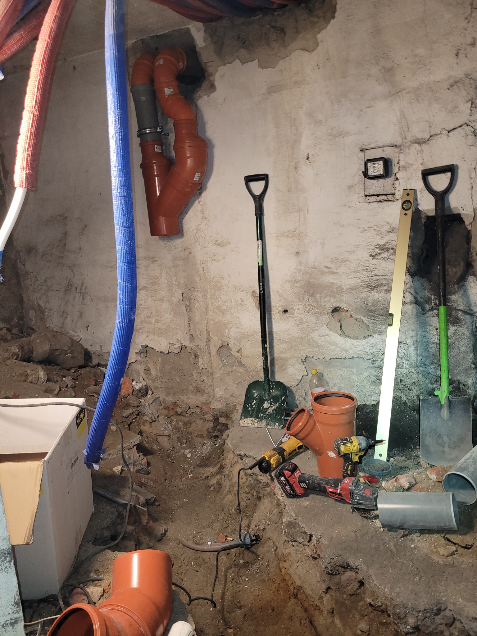 Montaż instalacji kanalizacyjnej z rur PCV w surowym wnętrzu. Widoczne narzędzia, poziomica i szpadle. Fragment instalacji w ścianie.