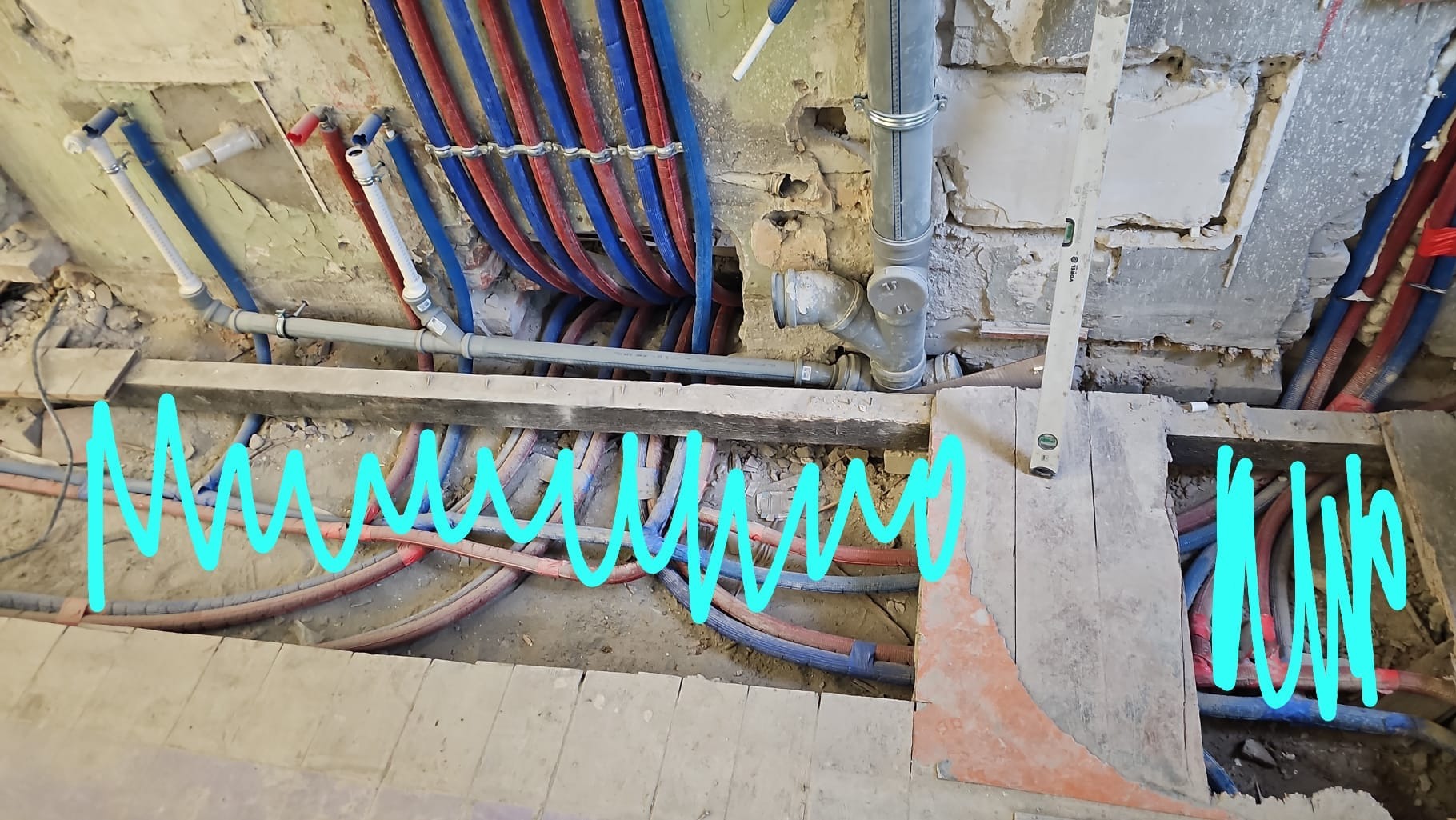 Instalacja wodna i kanalizacyjna w trakcie montażu, widoczne rury w kolorach niebieskim i czerwonym, poziomica. Prace instalacyjne w surowym stanie.