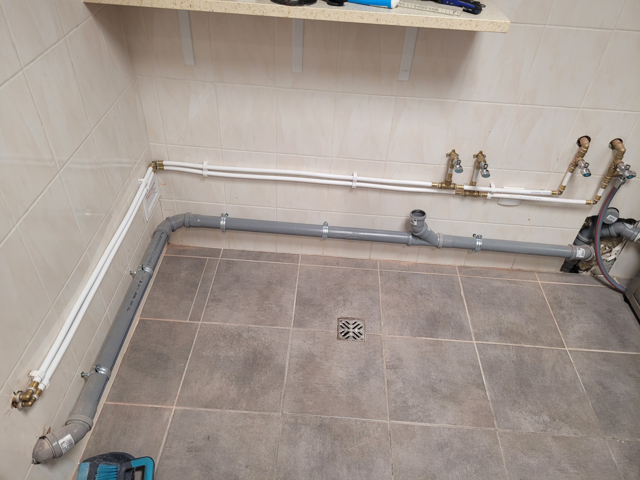 Szare i białe rury instalacji wodnej zamontowane w łazience z beżowymi płytkami, widoczny odpływ podłogowy i narzędzie. Instalacja po montażu.