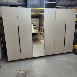 Firma Handlowo-Usługowa NATA Ryszard Kukla - Szafa z lustrem w garażu, szare fronty, czarne uchwyty, widoczne rury pod sufitem, betonowa podłoga, w lustrze odbija się warsztat.