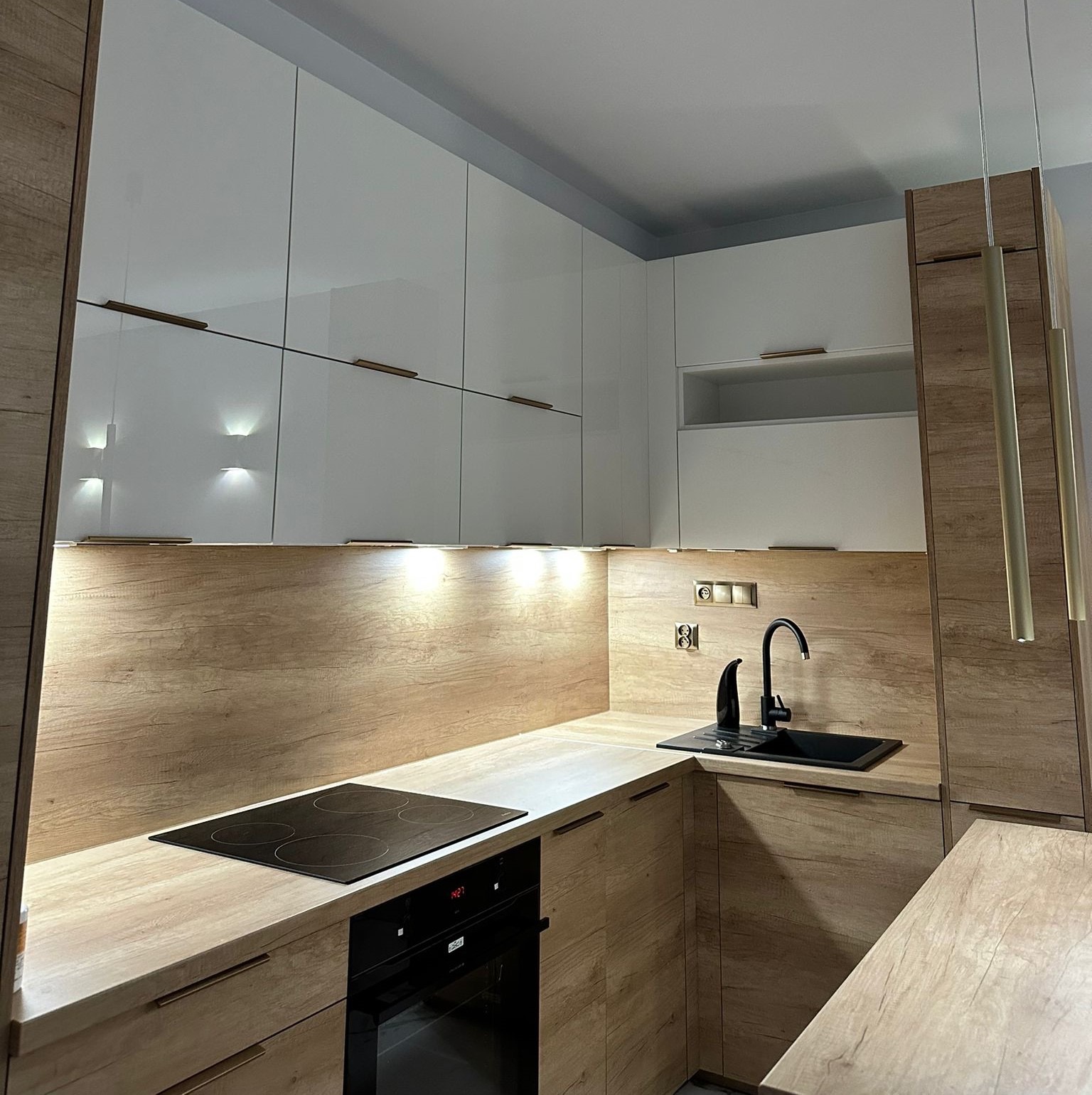 Nowoczesna kuchnia z białymi szafkami i drewnianymi akcentami. Czarny zlew i płyta indukcyjna. Oświetlenie podszafkowe i wiszące lampy nadają wnętrzu ciepły klimat.