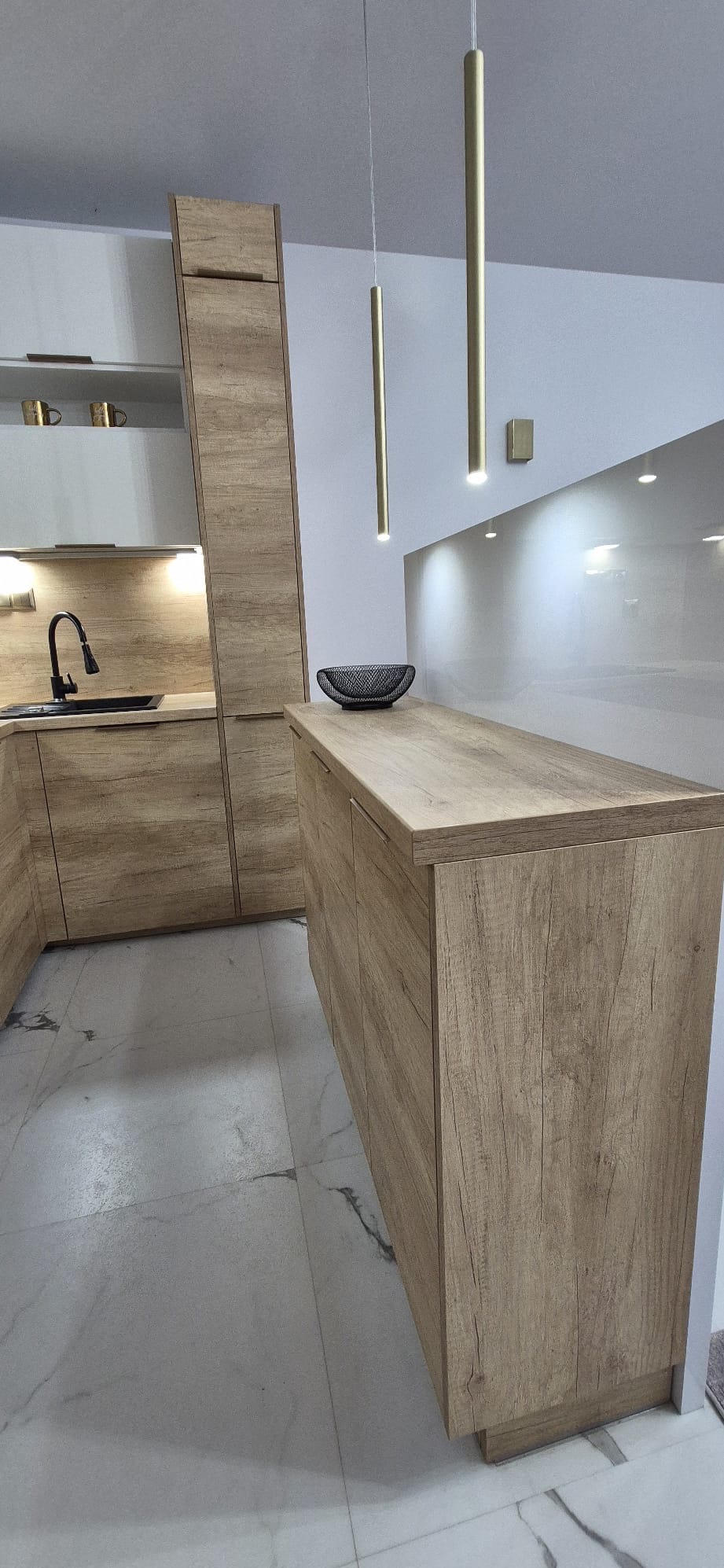 Nowoczesna kuchnia z drewnianymi frontami, minimalistycznym oświetleniem i marmurową podłogą. Na blacie dekoracyjna misa. Ujęcie z niskiego kąta.