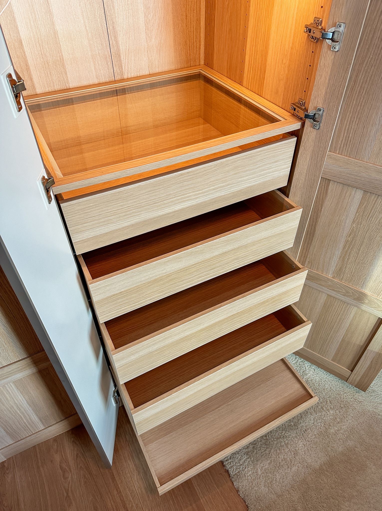 Otwarta szafa wnękowa z czterema wysuwanymi szufladami z jasnego drewna, widoczne zawiasy drzwi i podłoga z dywanem. Szafa wykonana na wymiar.
