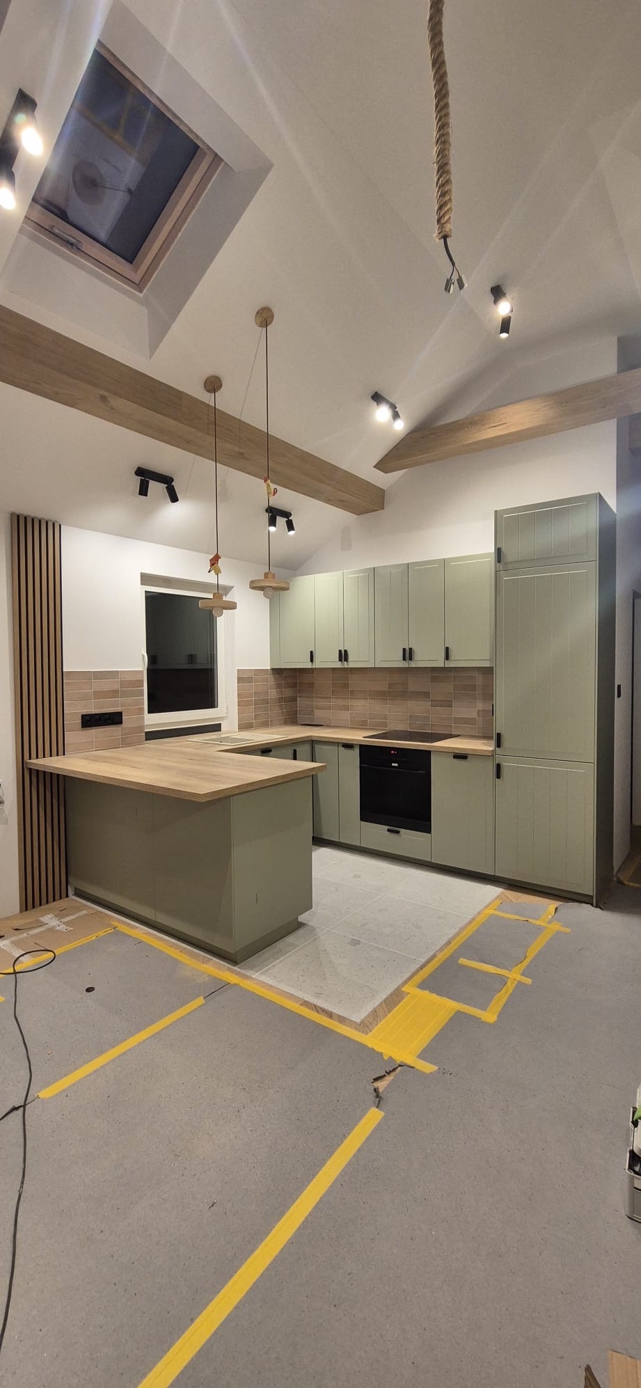 Jasna kuchnia w stylu rustykalnym z drewnianymi belkami stropowymi i wyspą. Widoczne niedokończone prace podłogowe z taśmą malarską. Oświetlenie punktowe i wiszące.