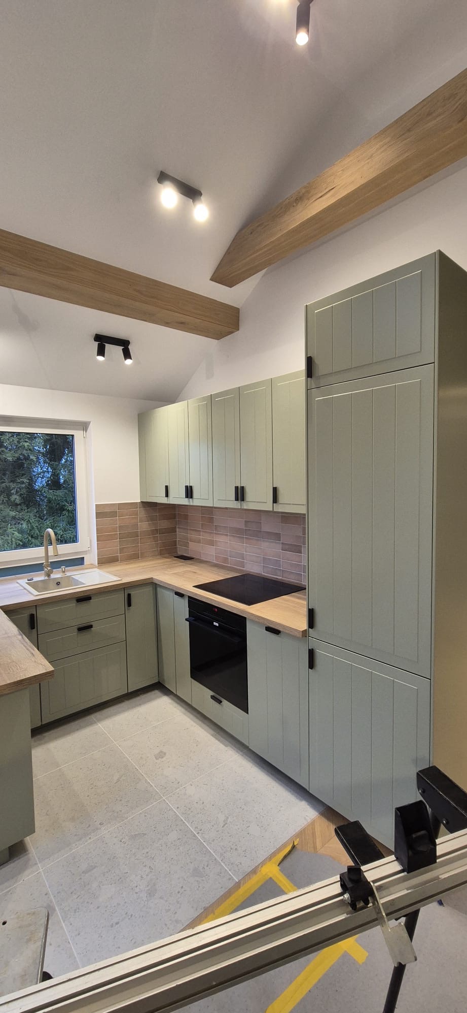 Jasnozielona kuchnia z drewnianymi blatami i beżową mozaiką nad blatem. Widoczne drewniane belki stropowe i narzędzia montażowe na podłodze.