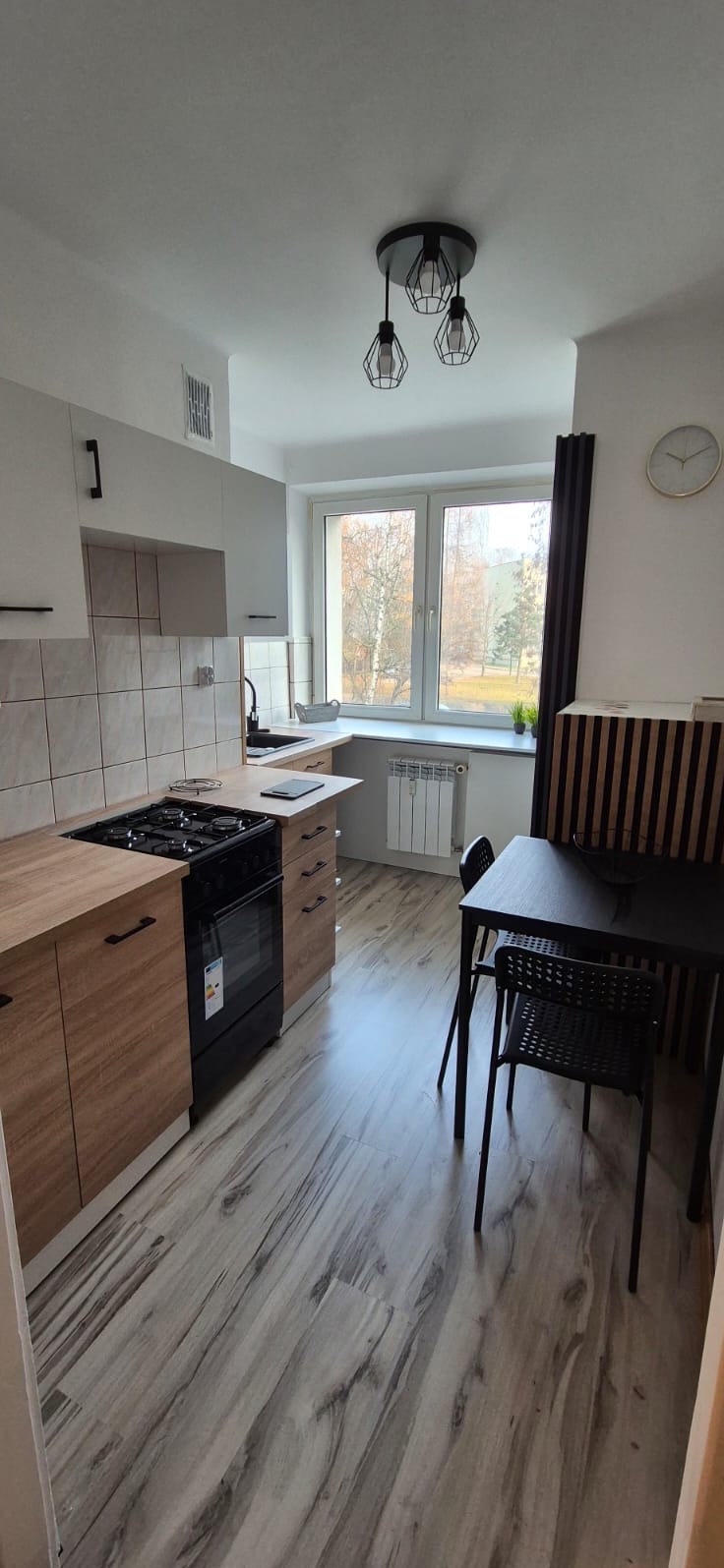 Jasna kuchnia z drewnianymi frontami, czarnym piekarnikiem i stołem. Na ścianie zegar, a przez okno widok na drzewa. Nowoczesne lampy wiszące.