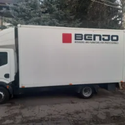 Biały samochód dostawczy z logo firmy BENJO Interiors and Furniture for Professionals zaparkowany na ulicy z widocznymi drzewami w tle.