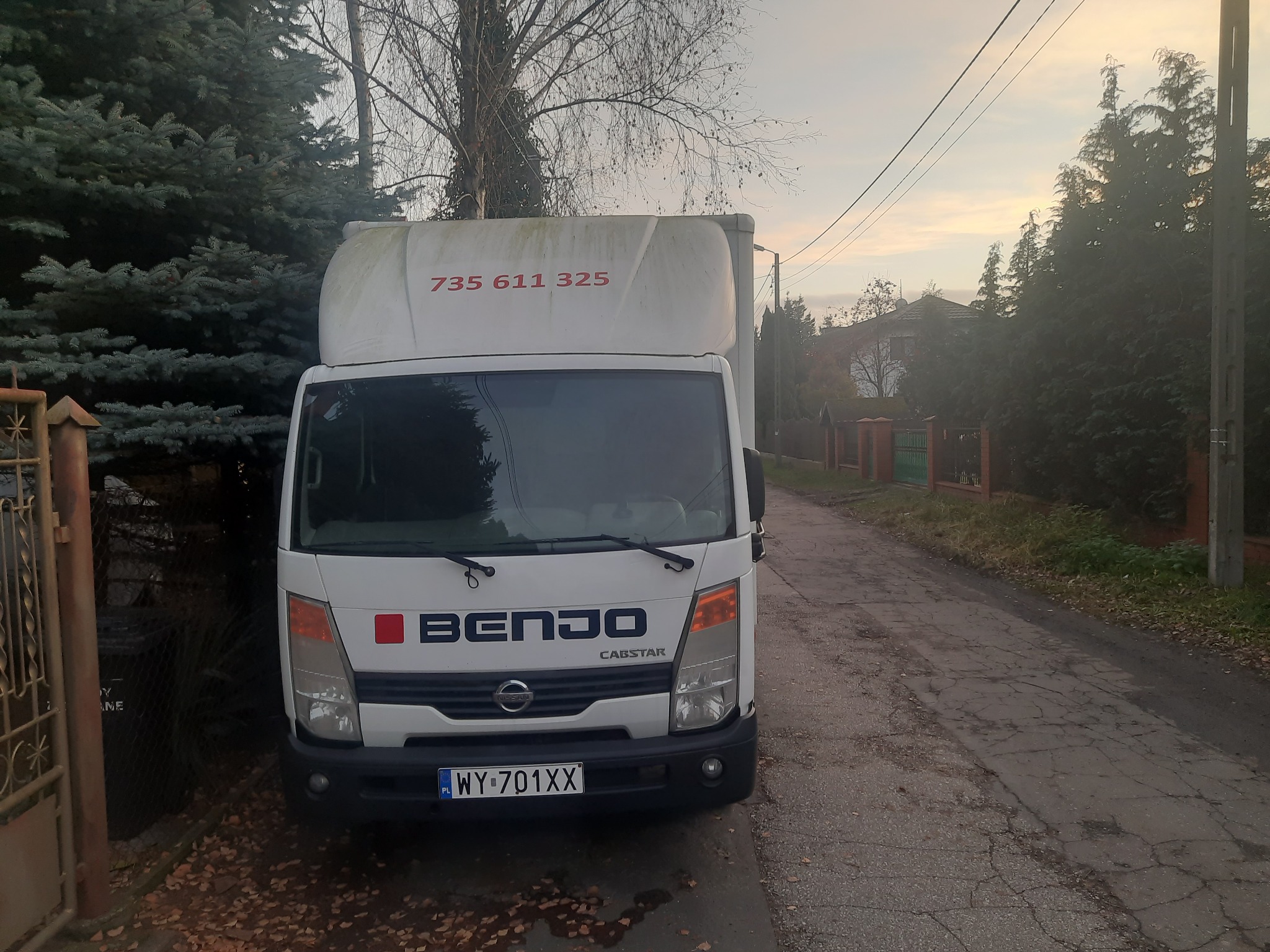 Biały samochód dostawczy marki Nissan Cabstar z numerem telefonu na nadwoziu, zaparkowany na pękniętej, asfaltowej drodze w jesiennej scenerii.