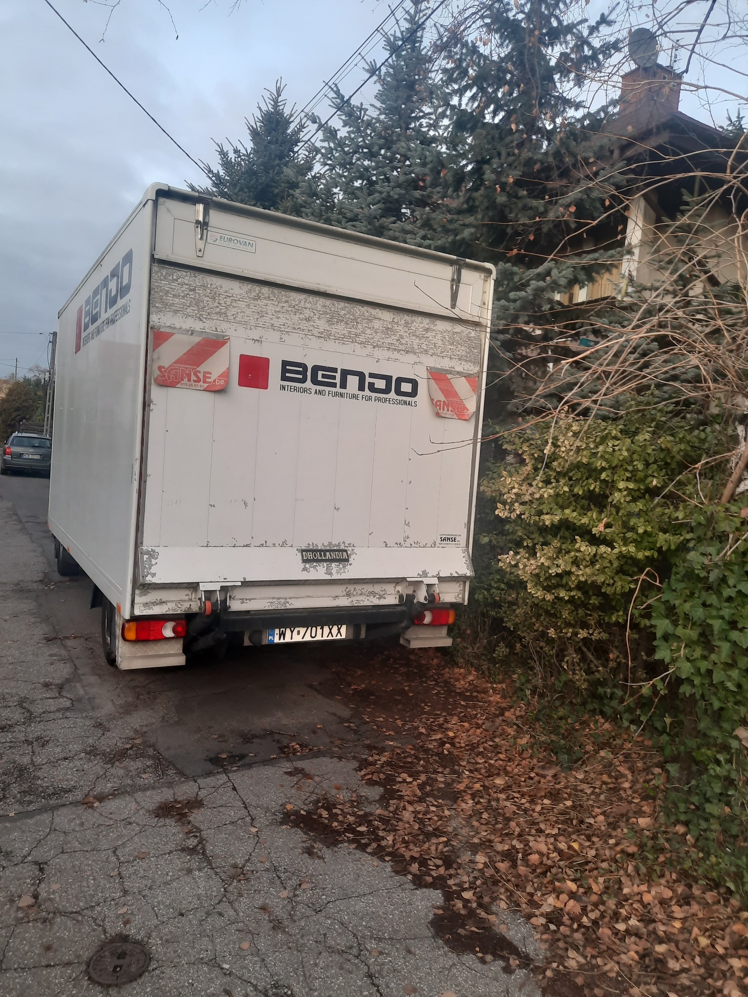 Tył białej furgonetki z logo 'BENJO' i napisem 'INTERIORS AND FURNITURE FOR PROFESSIONALS', zaparkowanej na ulicy w pobliżu zieleni i opadłych liści.