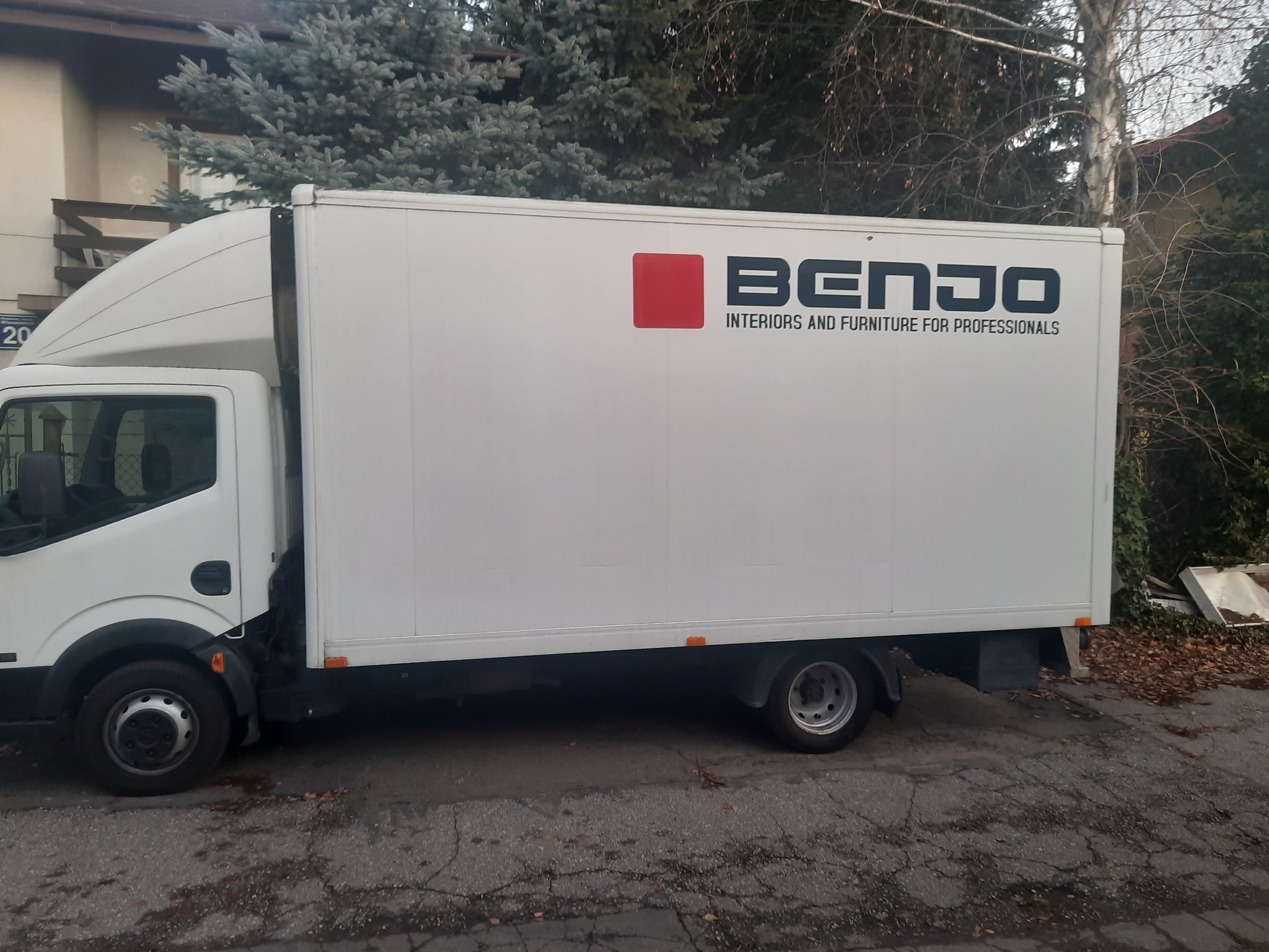 Biały samochód dostawczy z logo firmy BENJO Interiors and Furniture for Professionals zaparkowany na ulicy z widocznymi drzewami w tle.