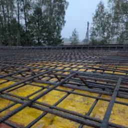 Siatka zbrojeniowa ułożona na żółtym szalunku, widok z bliska, w tle drzewa i pochmurne niebo.