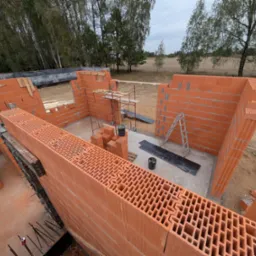 Wznoszenie ścian domu z czerwonej ceramiki poryzowanej, widoczne rusztowanie, drabina, zapas bloczków i narzędzia na placu budowy.