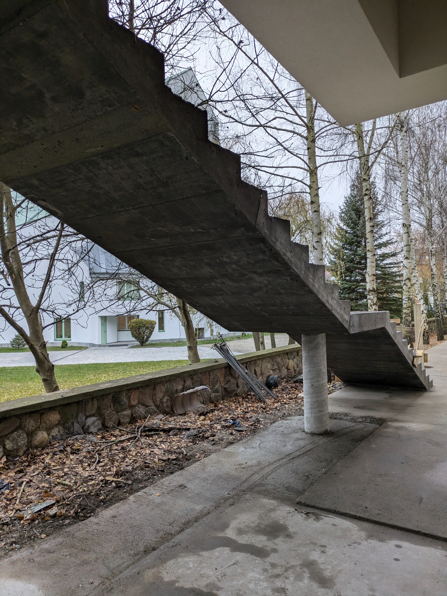 Surowe, betonowe schody zewnętrzne wsparte na kolumnie, widoczne na tle budynku i drzew bez liści, z rekwizytami budowlanymi i śladami po pracy na podłożu.