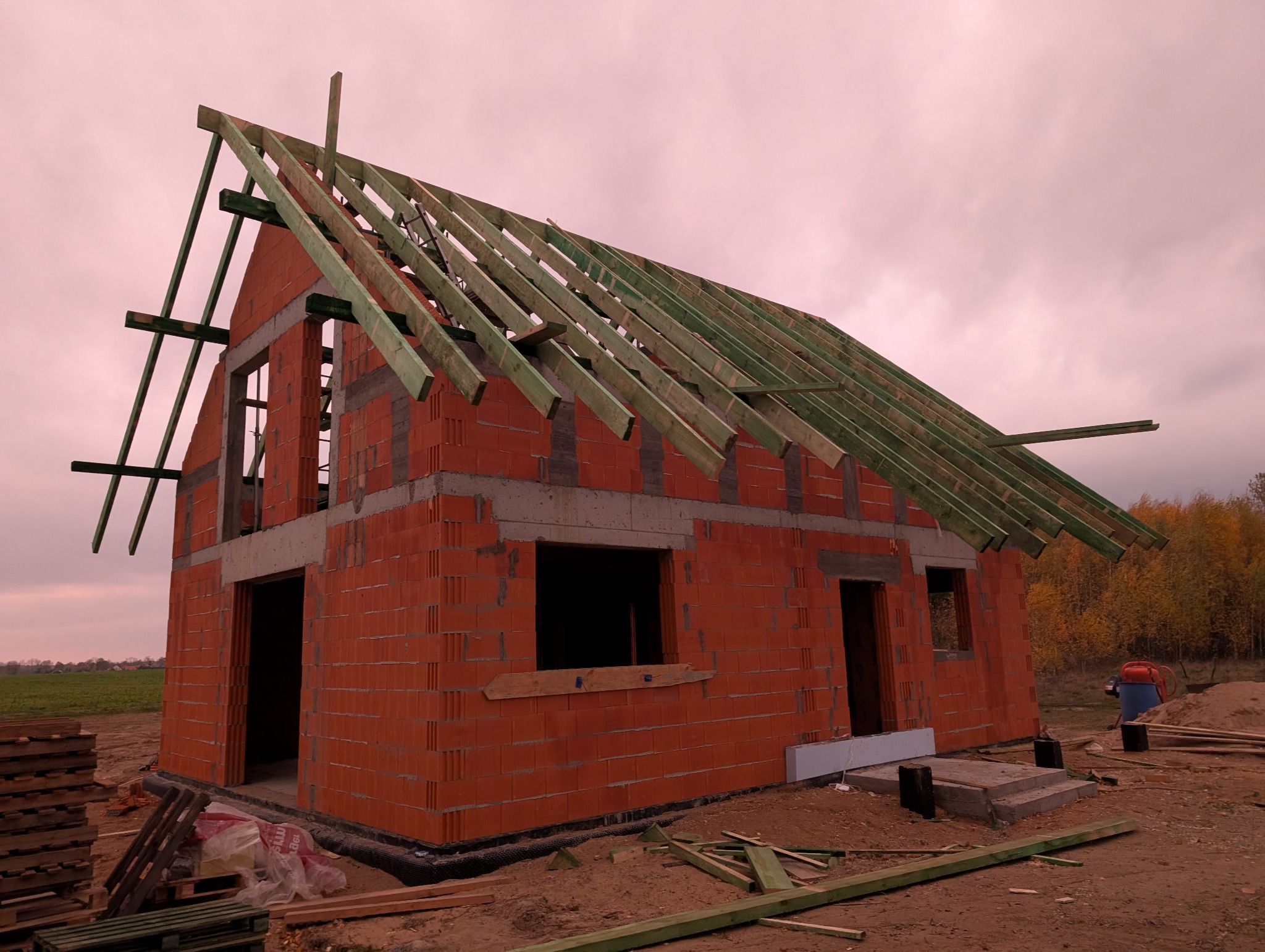 Surowy stan domu z czerwonej cegły z widoczną konstrukcją dachu z zielonych belek na tle pochmurnego nieba i jesiennego krajobrazu.
