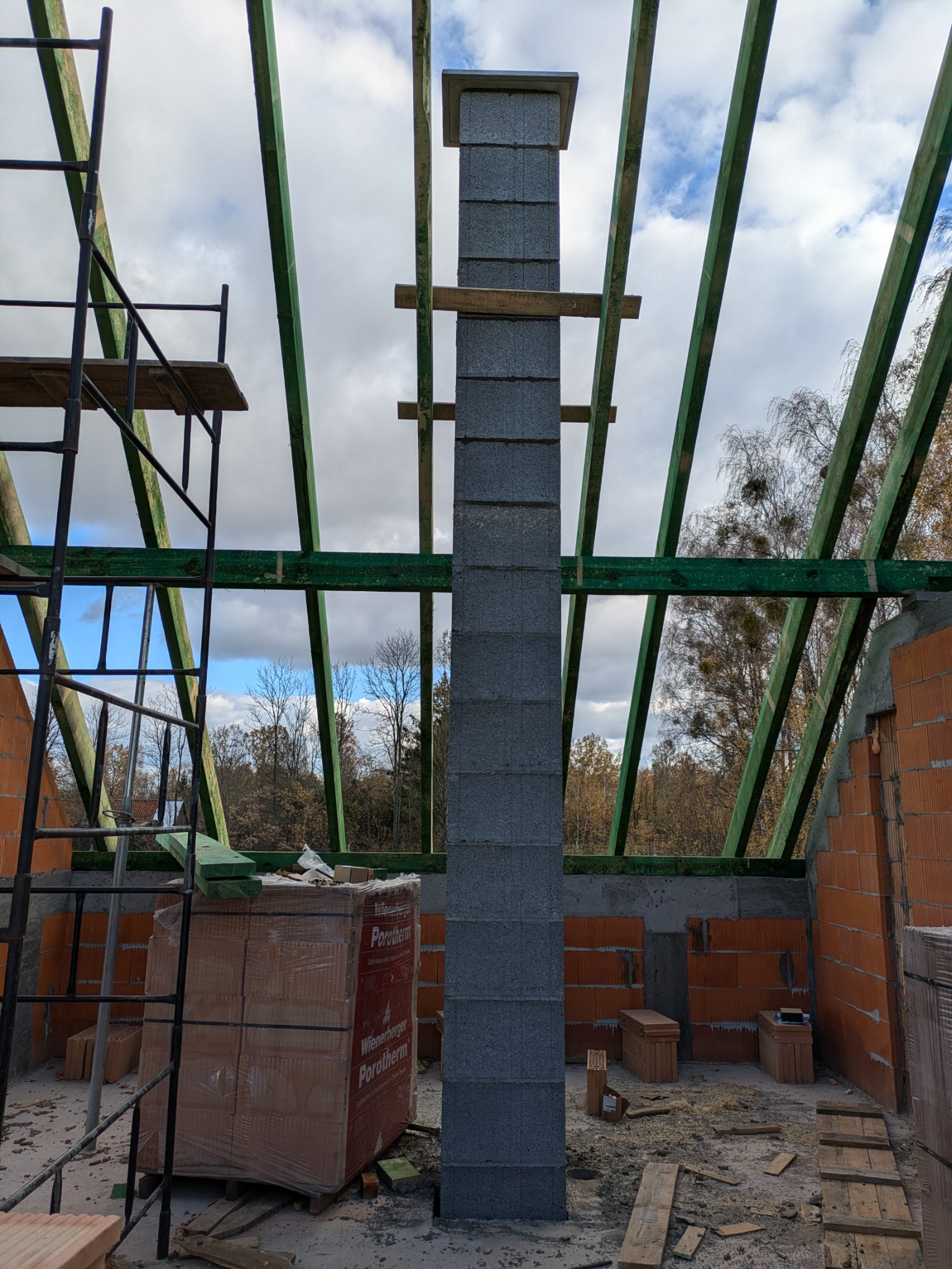 Wznoszenie komina z szarych bloczków kominowych na budowie domu, widoczne elementy konstrukcji dachu z zielonych belek i częściowo wymurowane ściany z czerwonej cegły Porotherm, w tle rusztowanie...