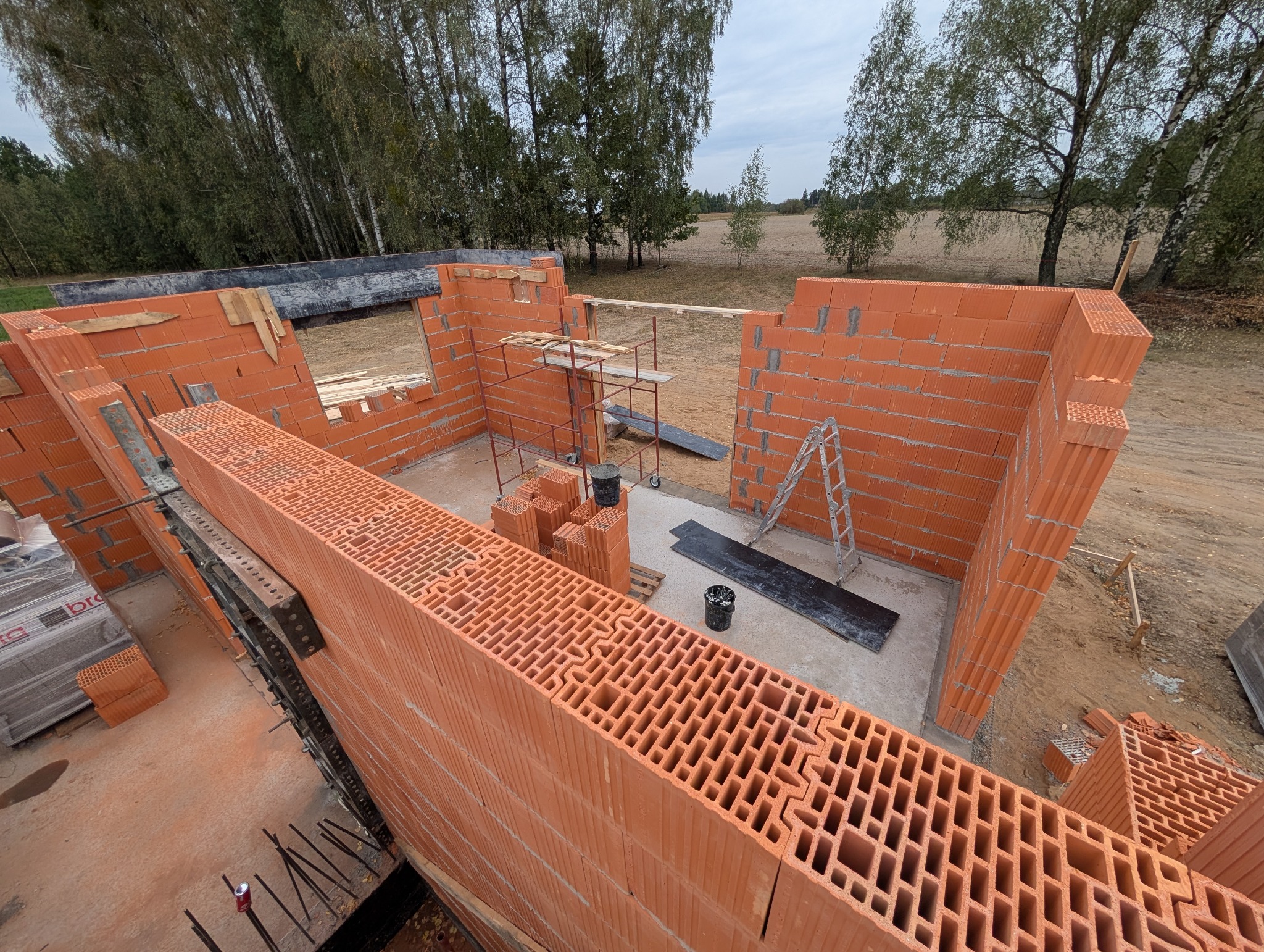 Wznoszenie ścian domu z czerwonej ceramiki poryzowanej, widoczne rusztowanie, drabina, zapas bloczków i narzędzia na placu budowy.